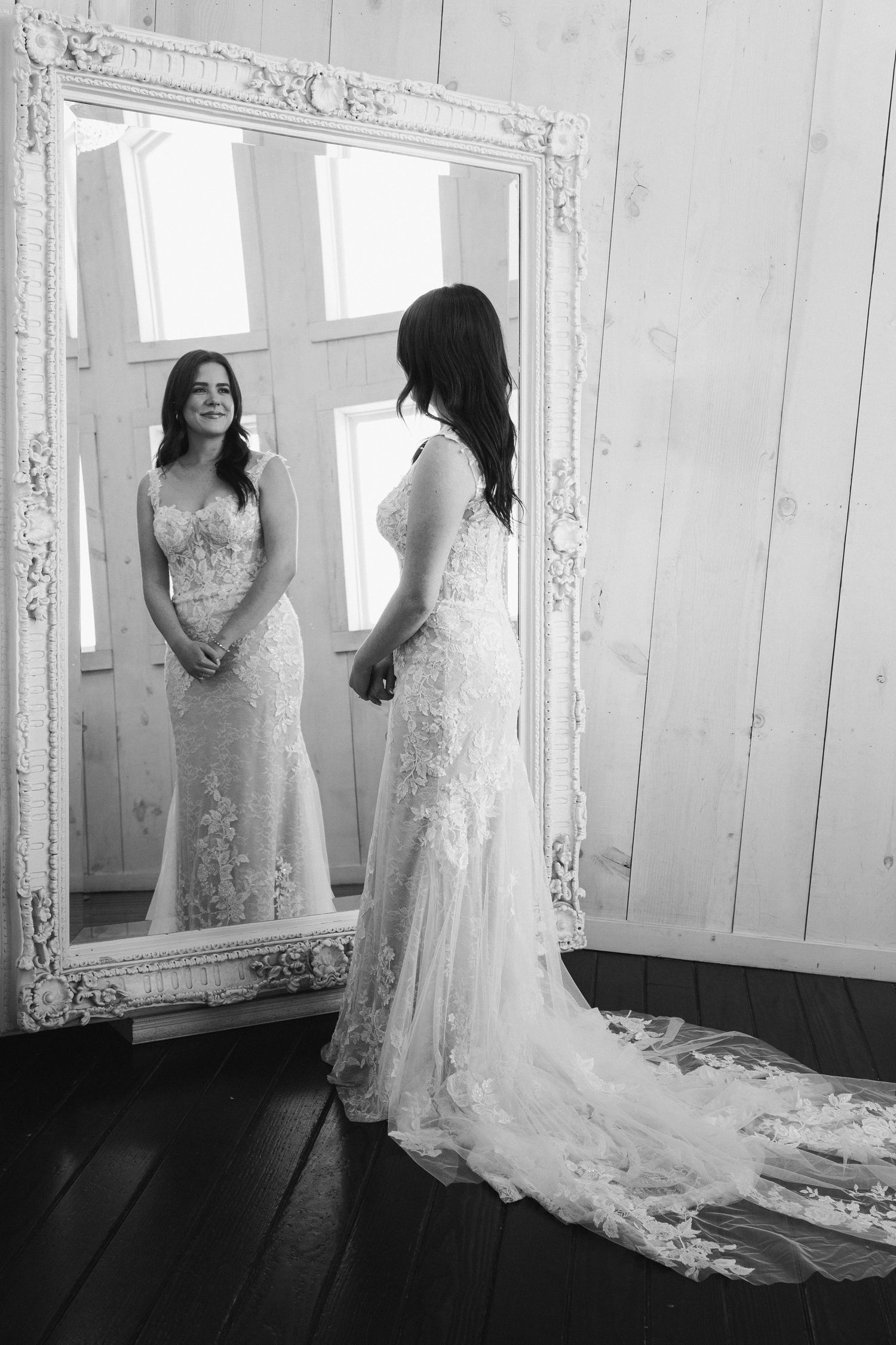 Bride in lace wedding gown admiring her reflection in ornate white mirror in black and white photo.