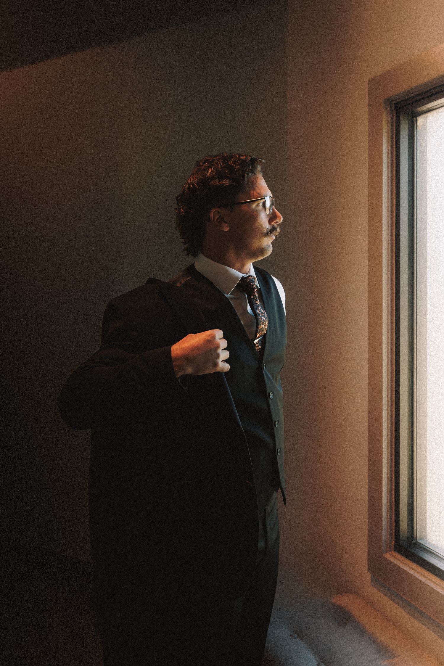 Dapper groom in black suit and glasses looks out window in dramatic moody lighting.