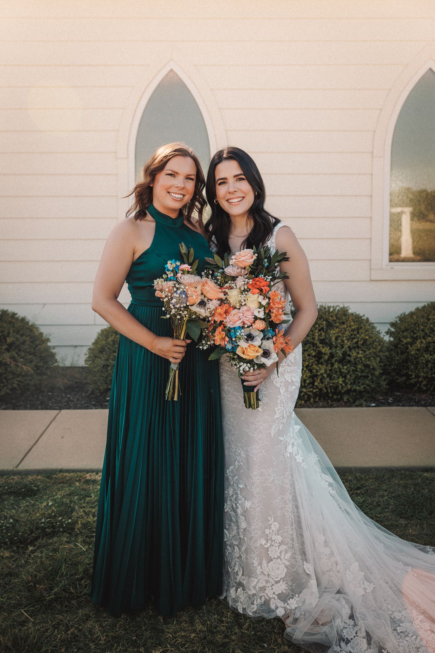 Bride in fitted lace wedding gown poses with bridesmaid in green halter dress holding bouquets.