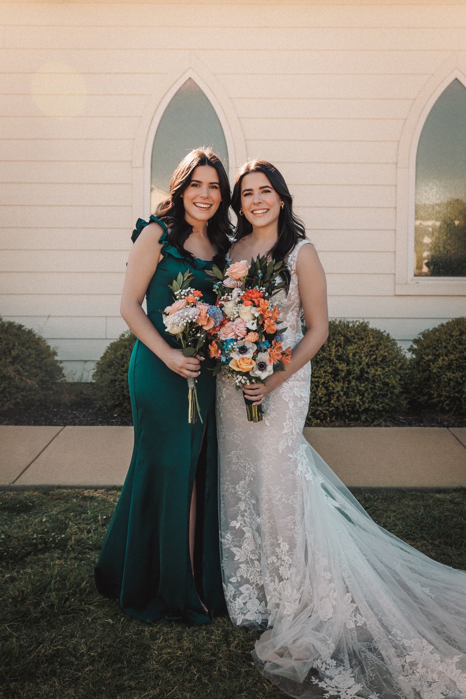 Bride in lace gown and bridesmaid in emerald dress hold colorful bouquets outside white chapel.