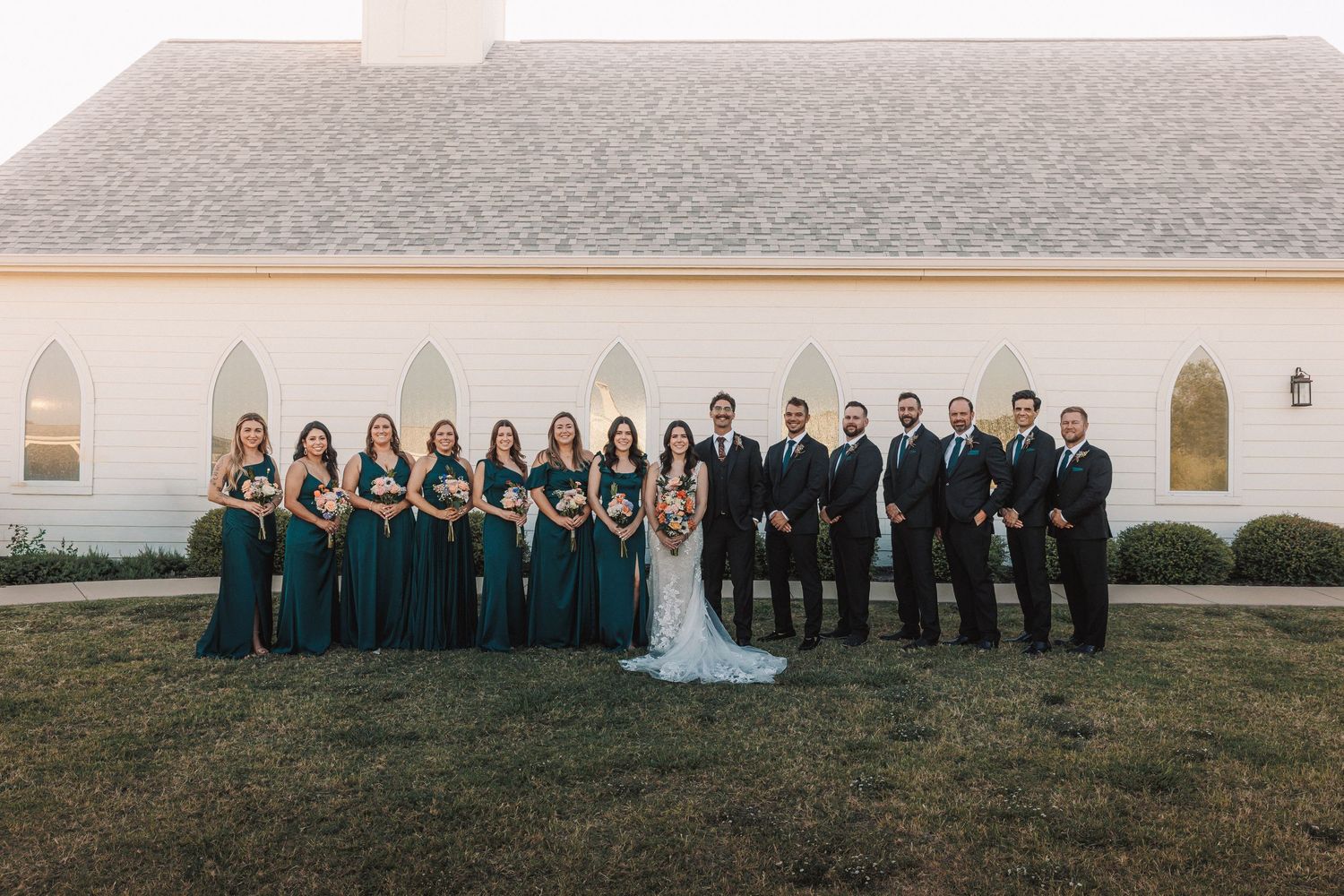 Wedding party poses outside white chapel, bridesmaids in emerald gowns and groomsmen in dark suits.