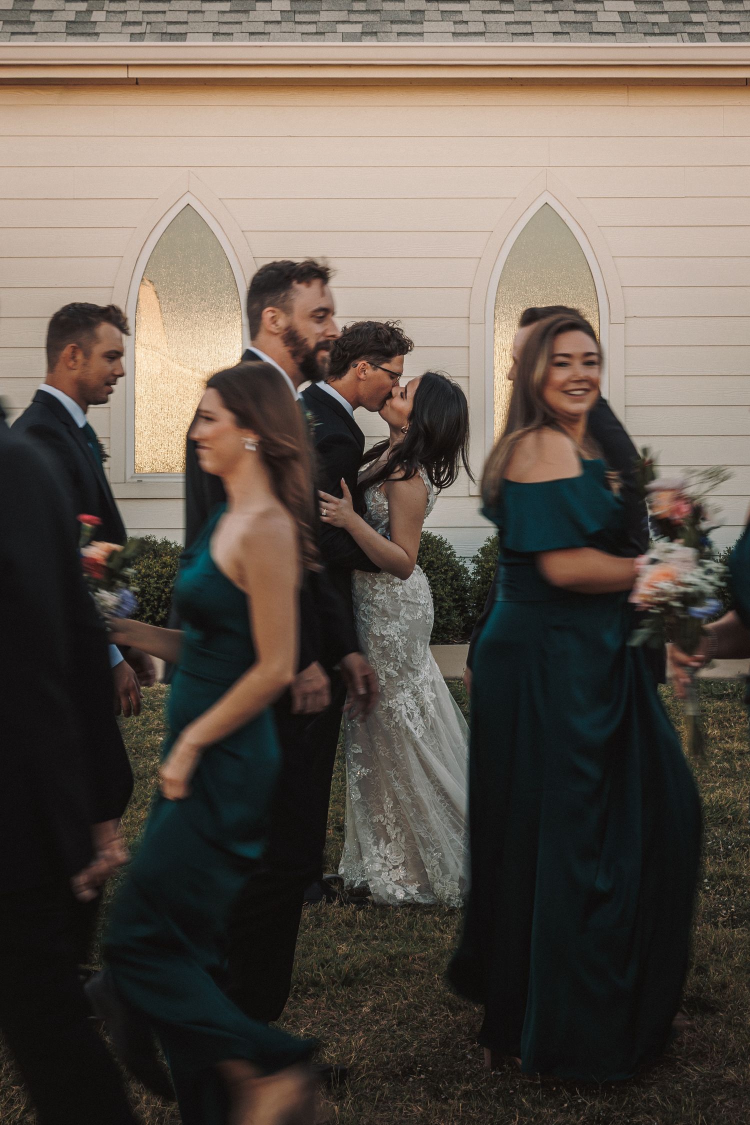 Newlyweds share a kiss surrounded by bridesmaids in emerald gowns and groomsmen outside a white chapel.