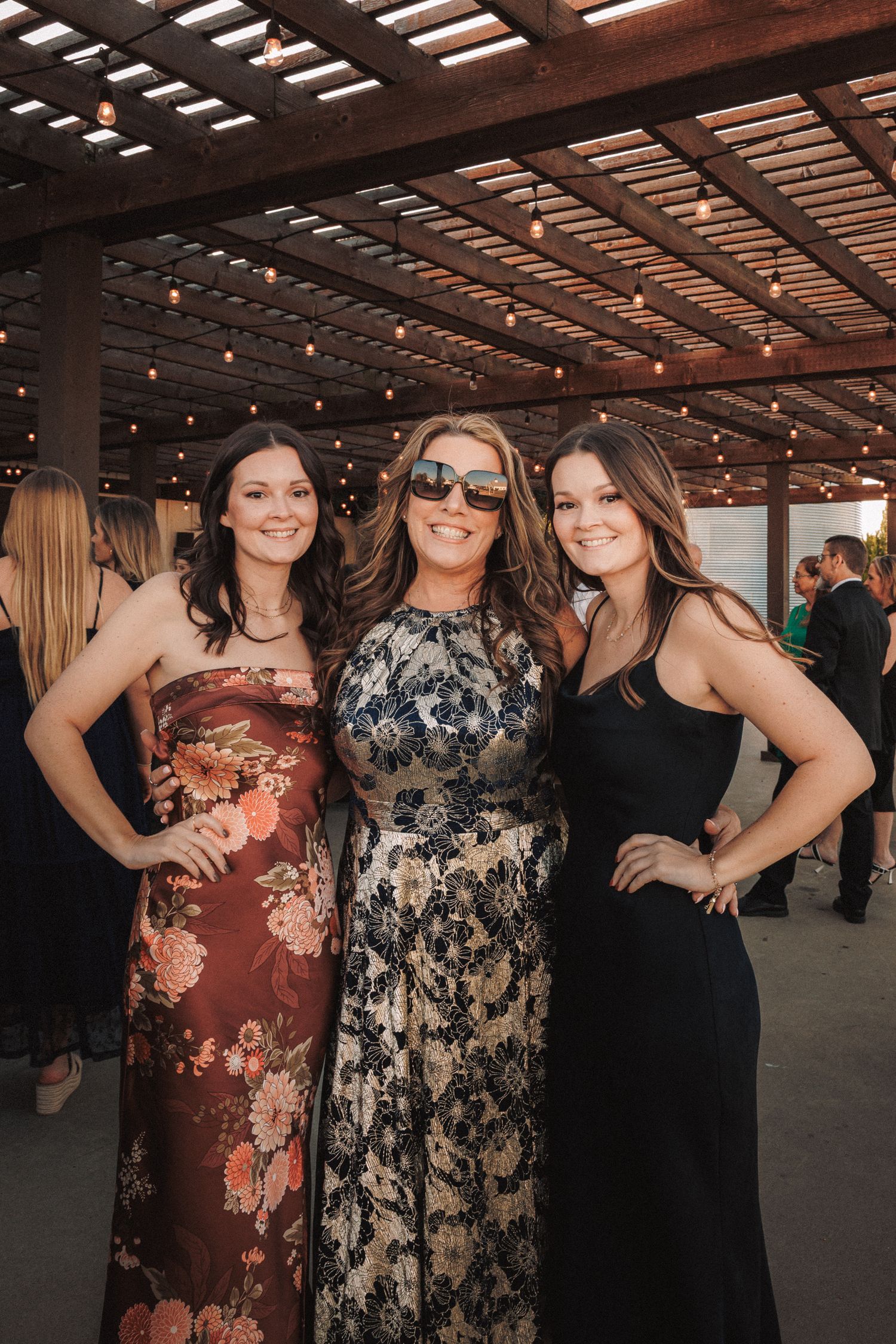 Three women in elegant formal dresses posing together at outdoor wedding reception.