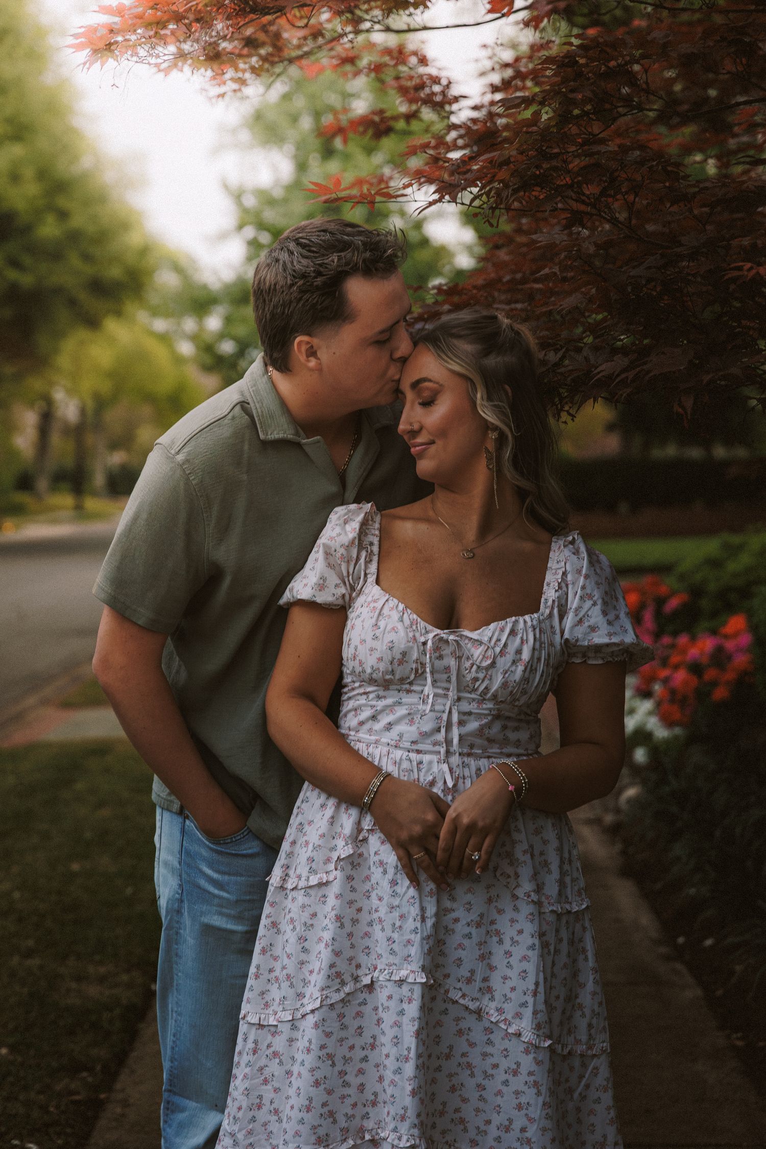 A man kisses a woman on the cheek as she smiles in a floral dress outdoors.