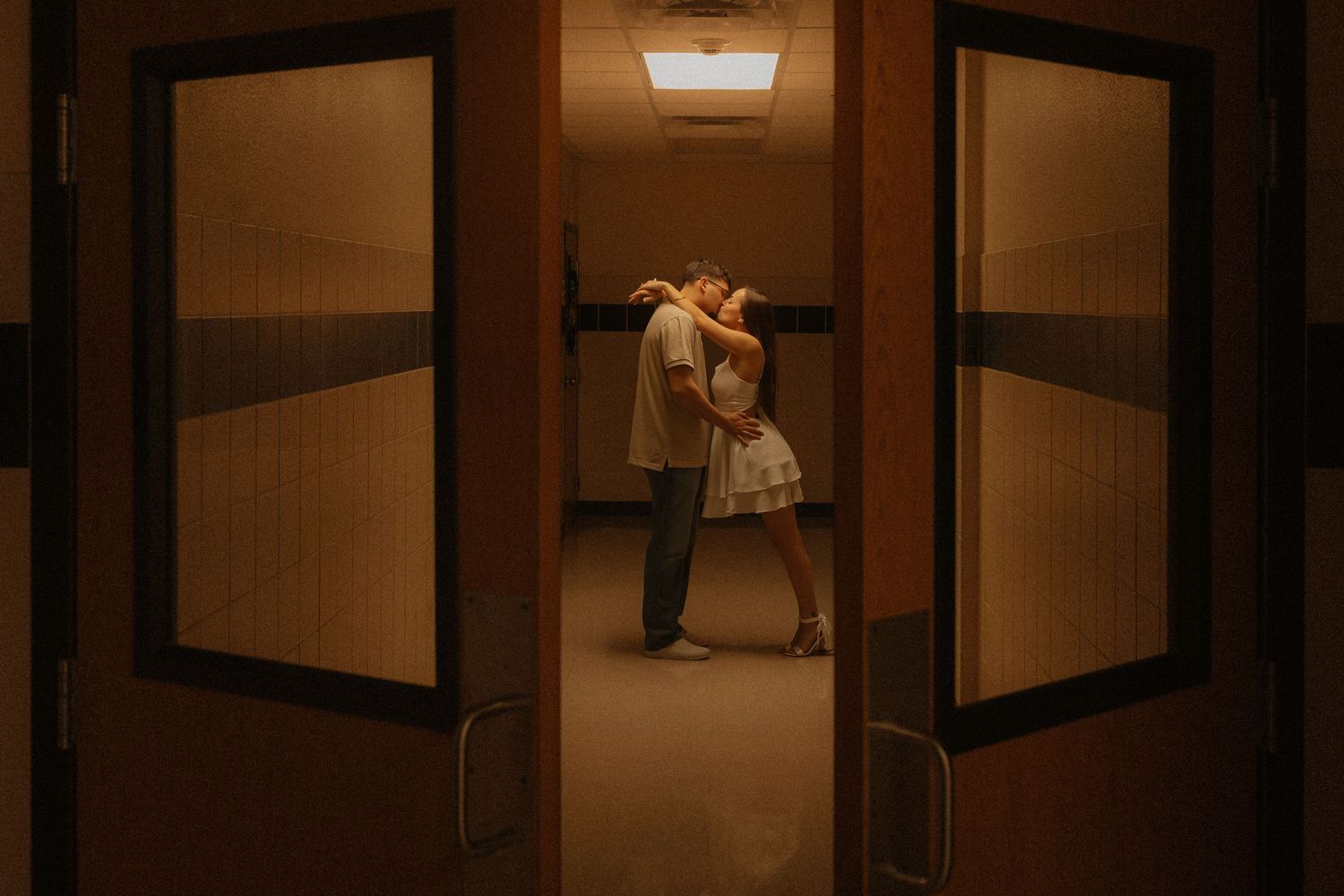 Couple sharing a romantic embrace in a warmly lit hallway, framed by wooden doors in a cinematic setting.