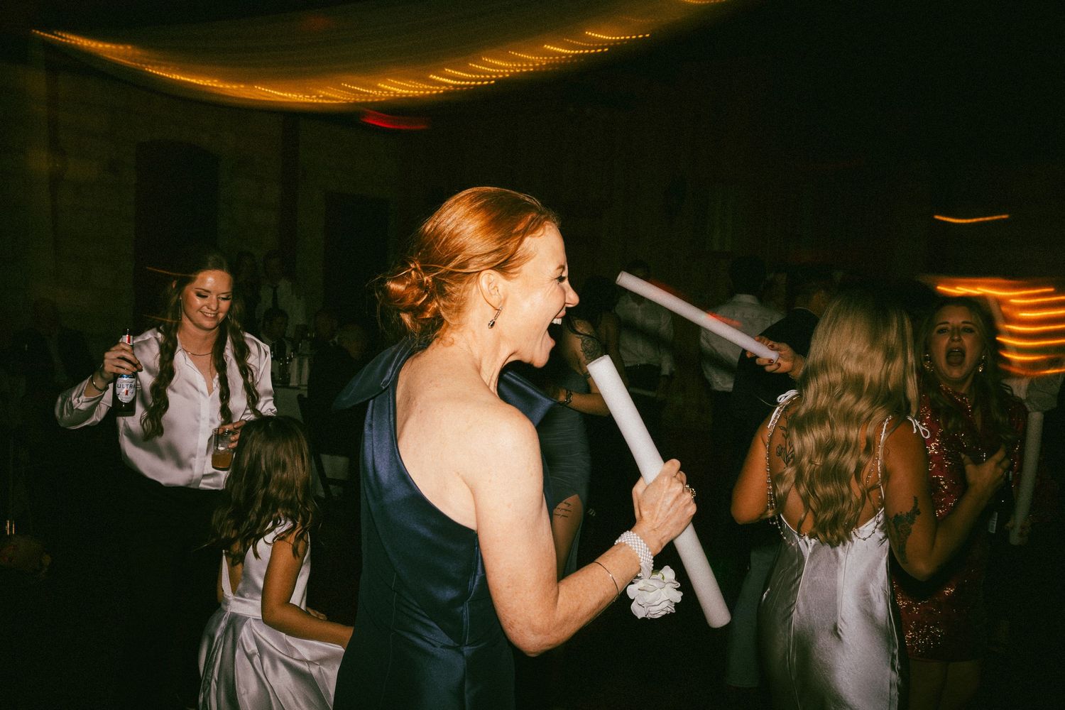 Woman in navy dress laughing while holding glow stick on wedding reception dance floor.