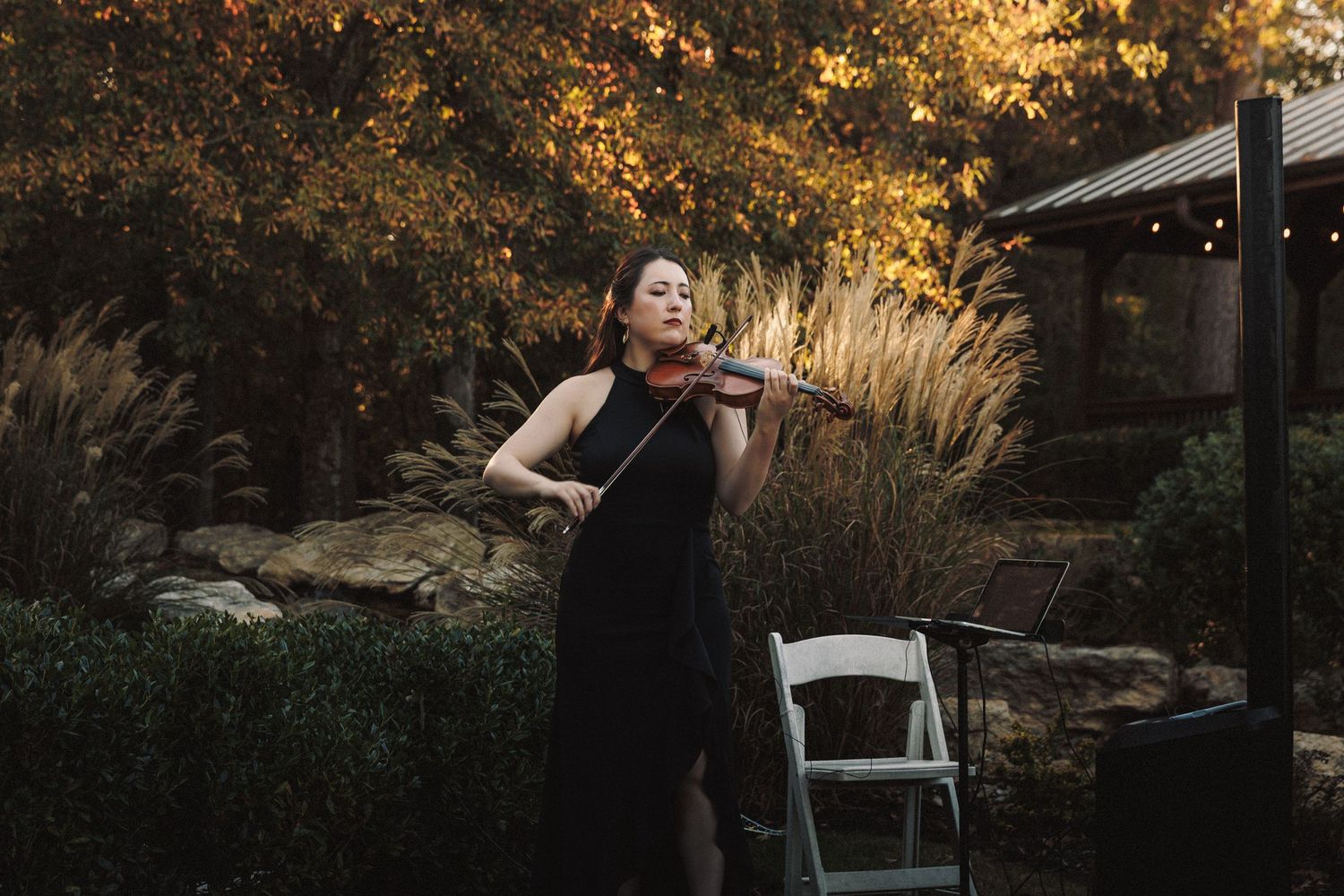 Violinist in black dress performing outdoors at wedding ceremony amid autumn foliage.