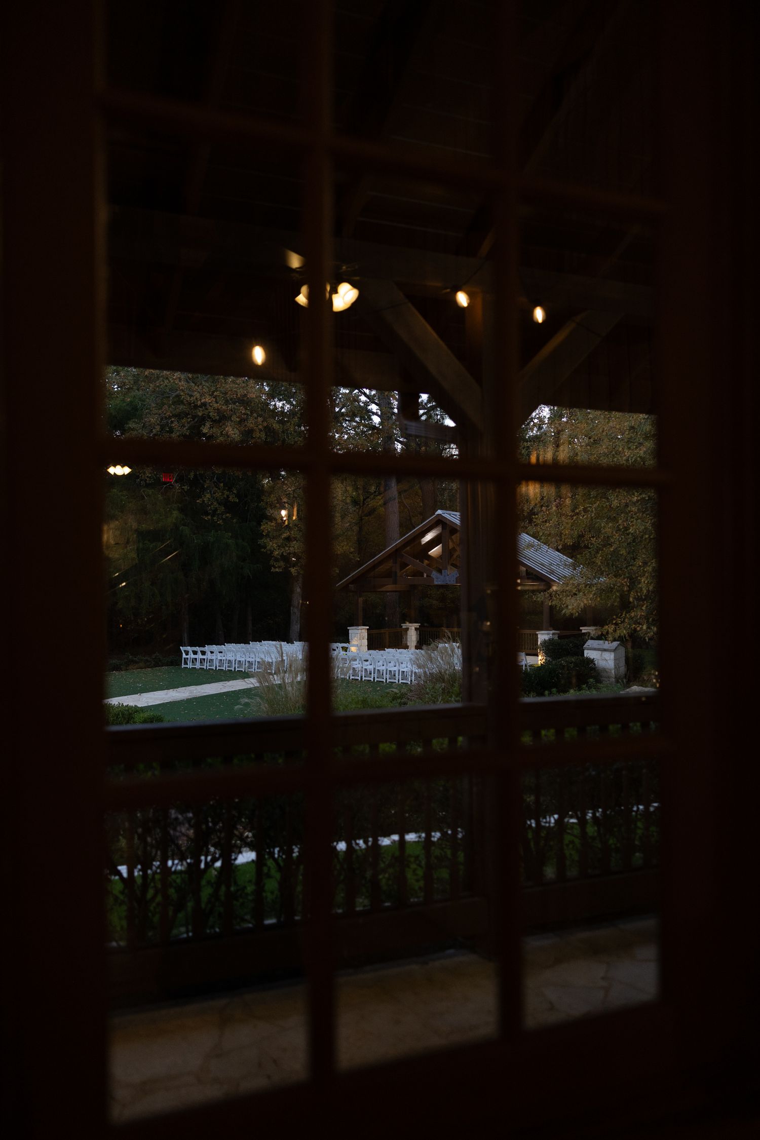 View through a window of an outdoor wedding ceremony setup with string lights and white chairs.