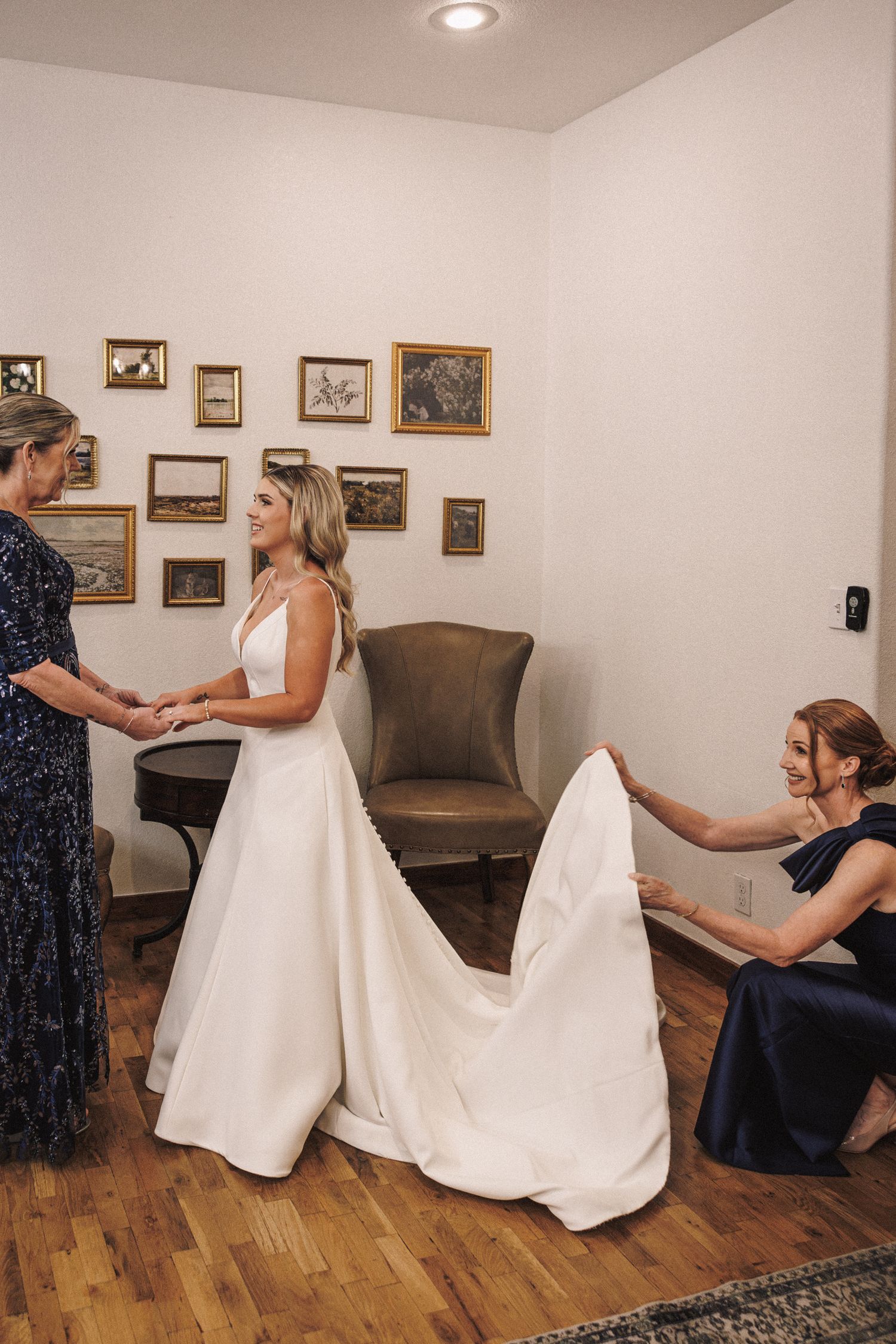 Bride in white gown assisted by two women adjusting her dress train in a warmly lit room with framed art.