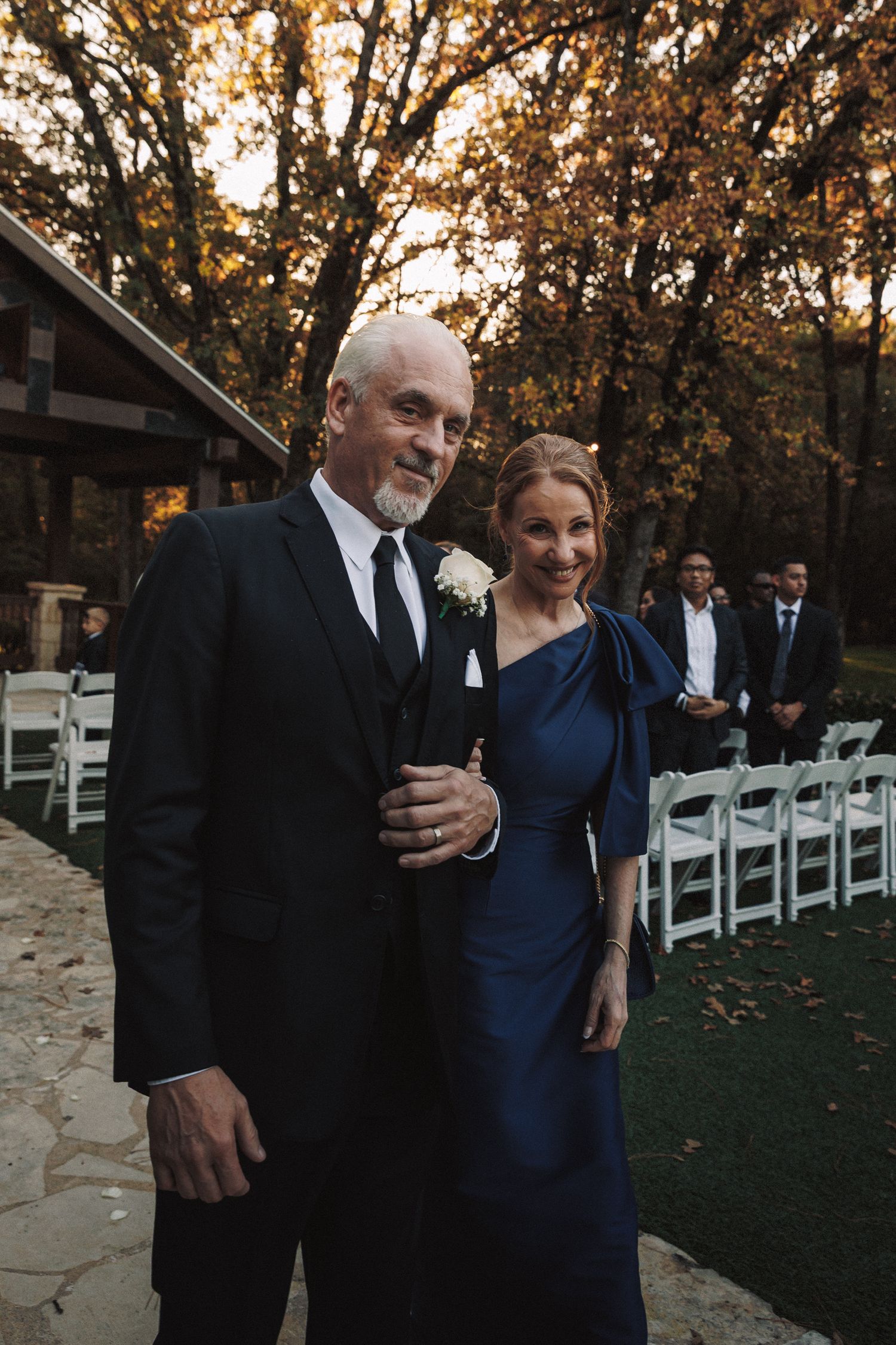 Elegant couple in formal attire posing near ceremony chairs in autumn outdoor setting.