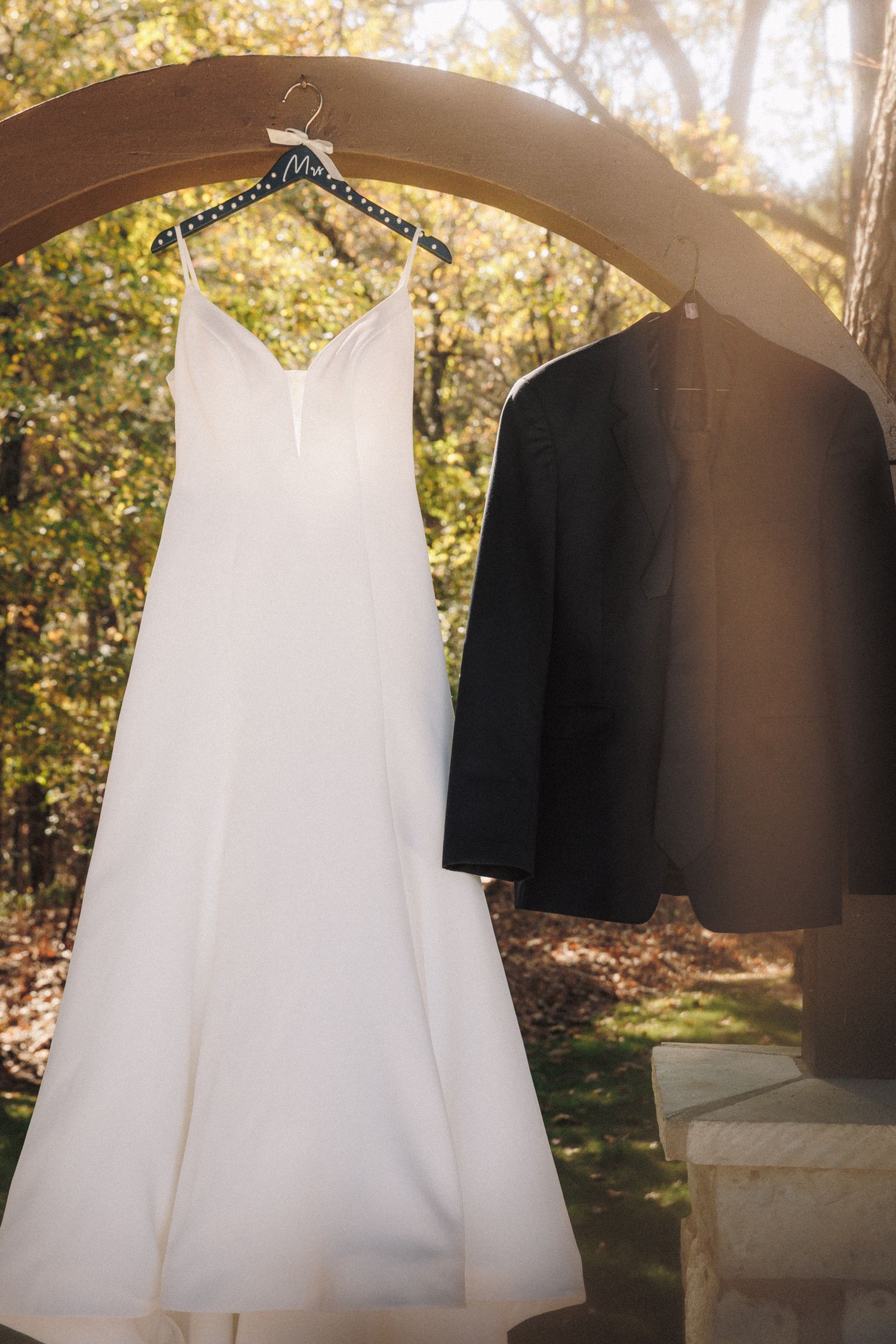 White wedding dress and black tuxedo jacket hanging on wooden arch outdoors with autumn trees in background.