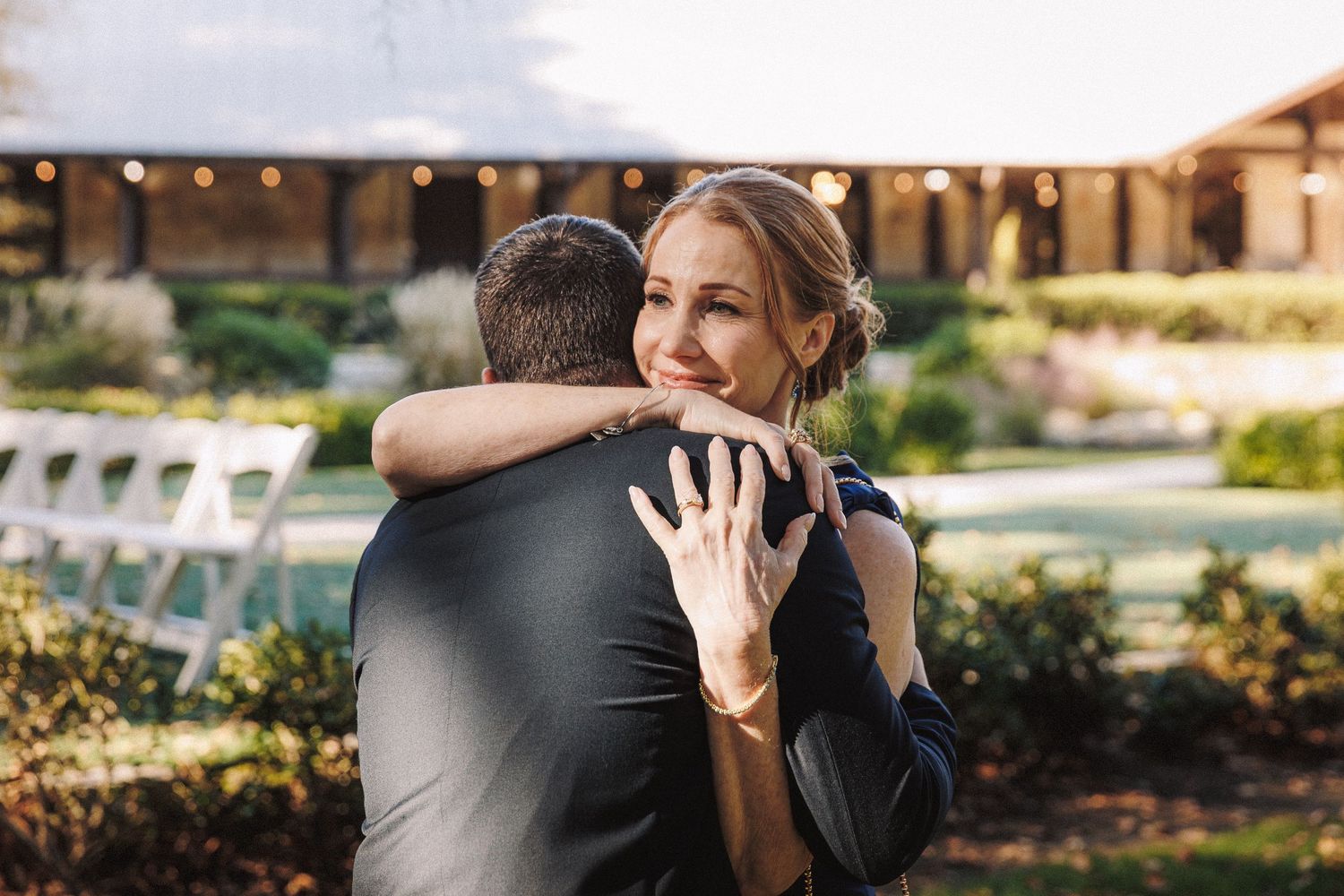 Emotional bride with updo hugging groom outdoors at elegant garden wedding venue with string lights.