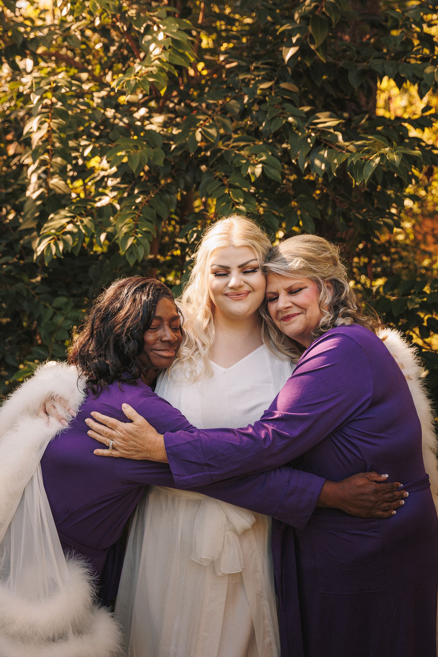 Bride in white lace gown shares emotional embrace with two women in purple dresses outdoors.