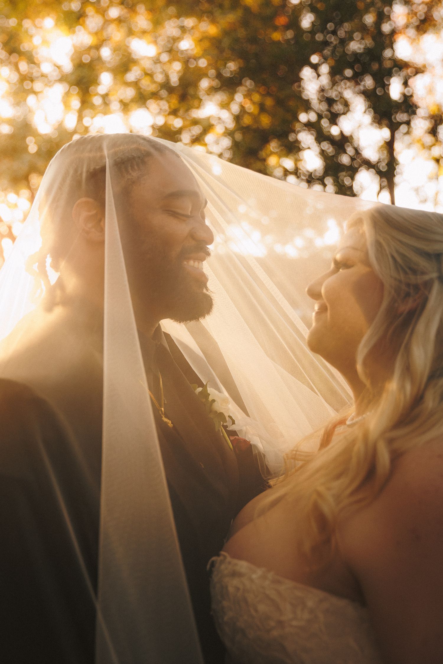 Bride and groom sharing an intimate moment under a wedding veil with golden sunlight filtering through autumn trees.