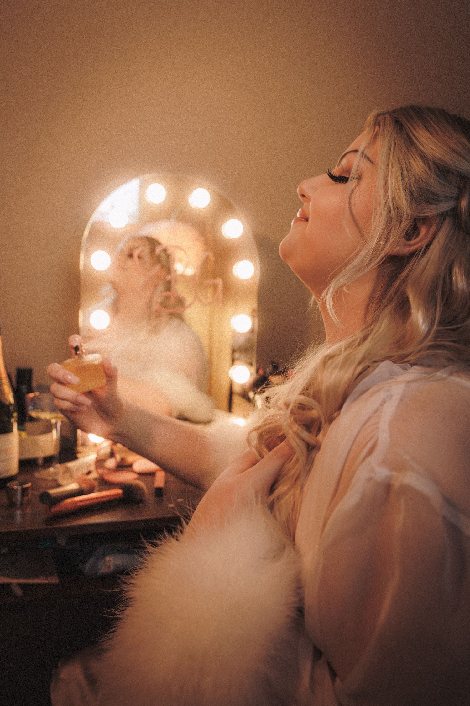 Bride in white robe spraying perfume at lit vanity mirror with warm Hollywood bulbs.