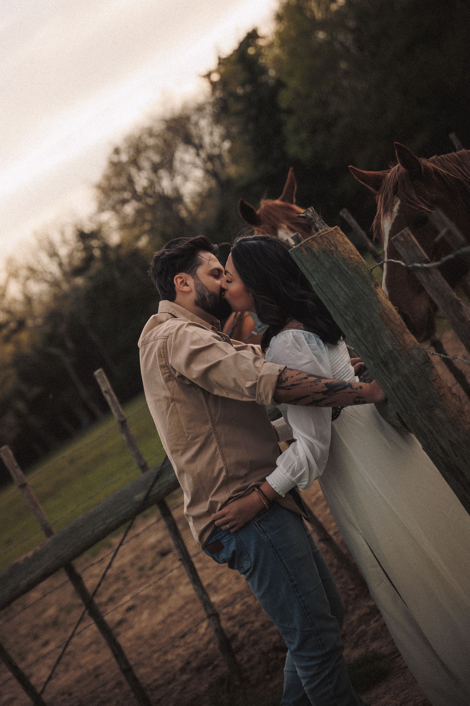 Couple kissing tenderly by a wooden fence with horses grazing in a scenic countryside setting.