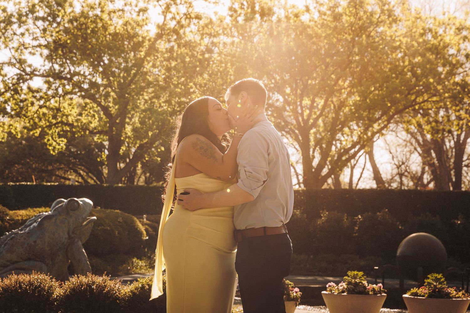Couple sharing a romantic kiss at golden hour in a sunlit garden, woman in yellow dress embraced by man in blue shirt.