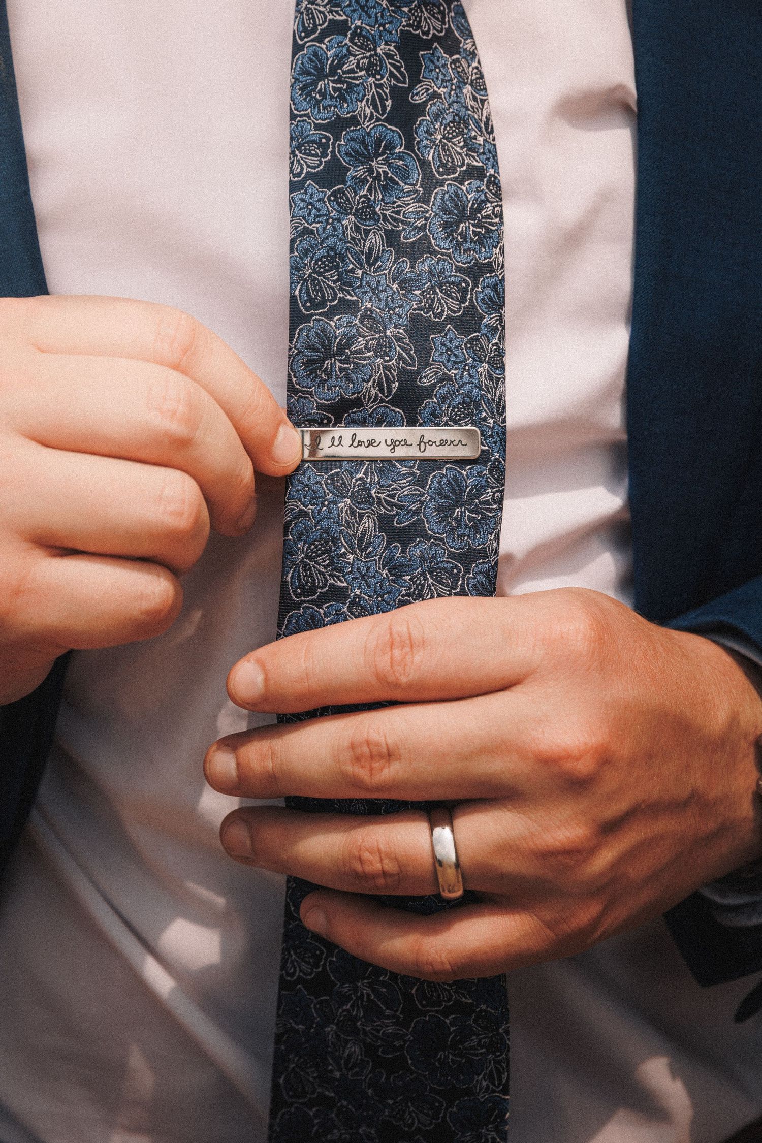 Close-up of hands adjusting a floral patterned blue tie with a tie clip and wedding ring.