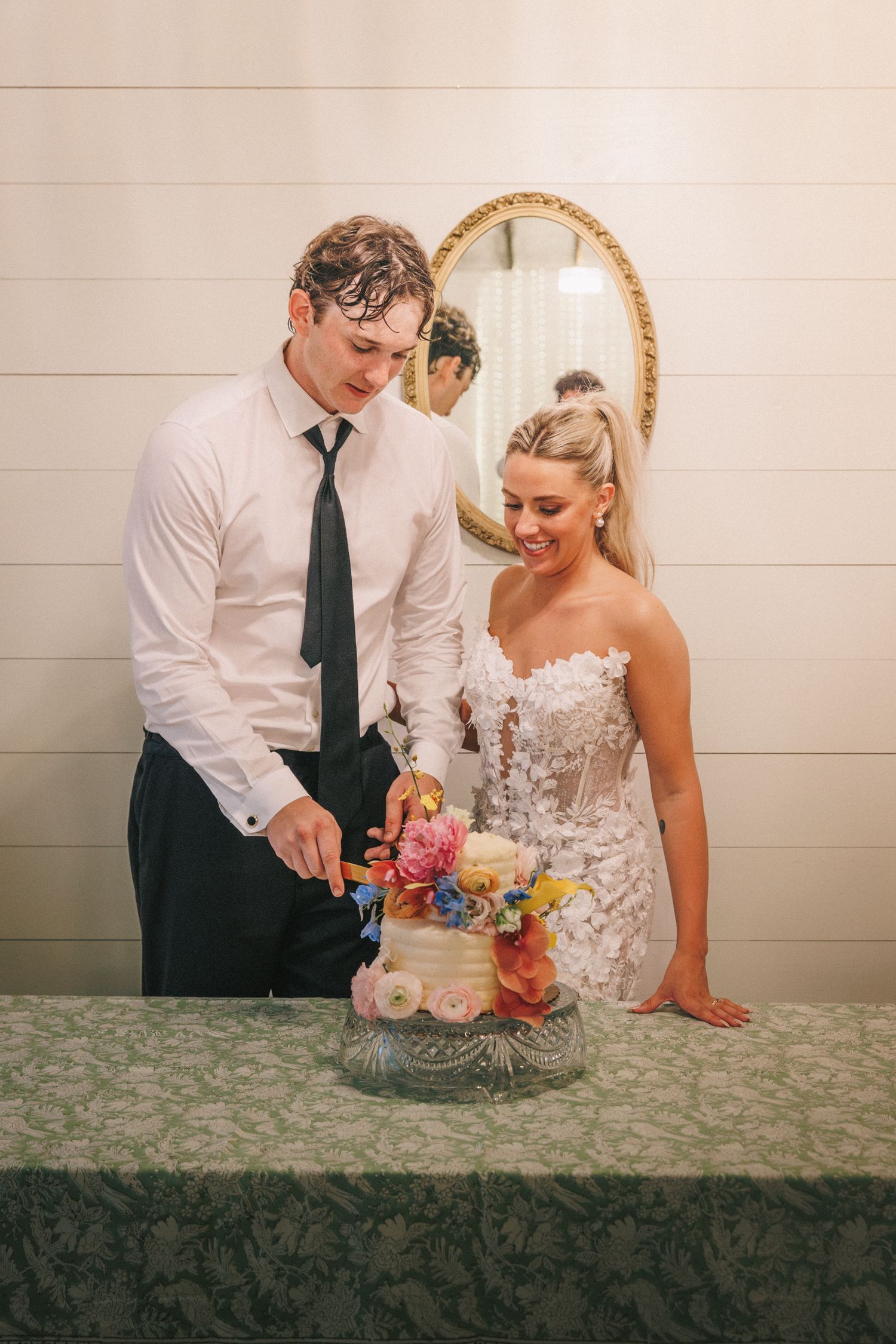 A couple cuts their colorful floral wedding cake together in front of an ornate gold mirror.