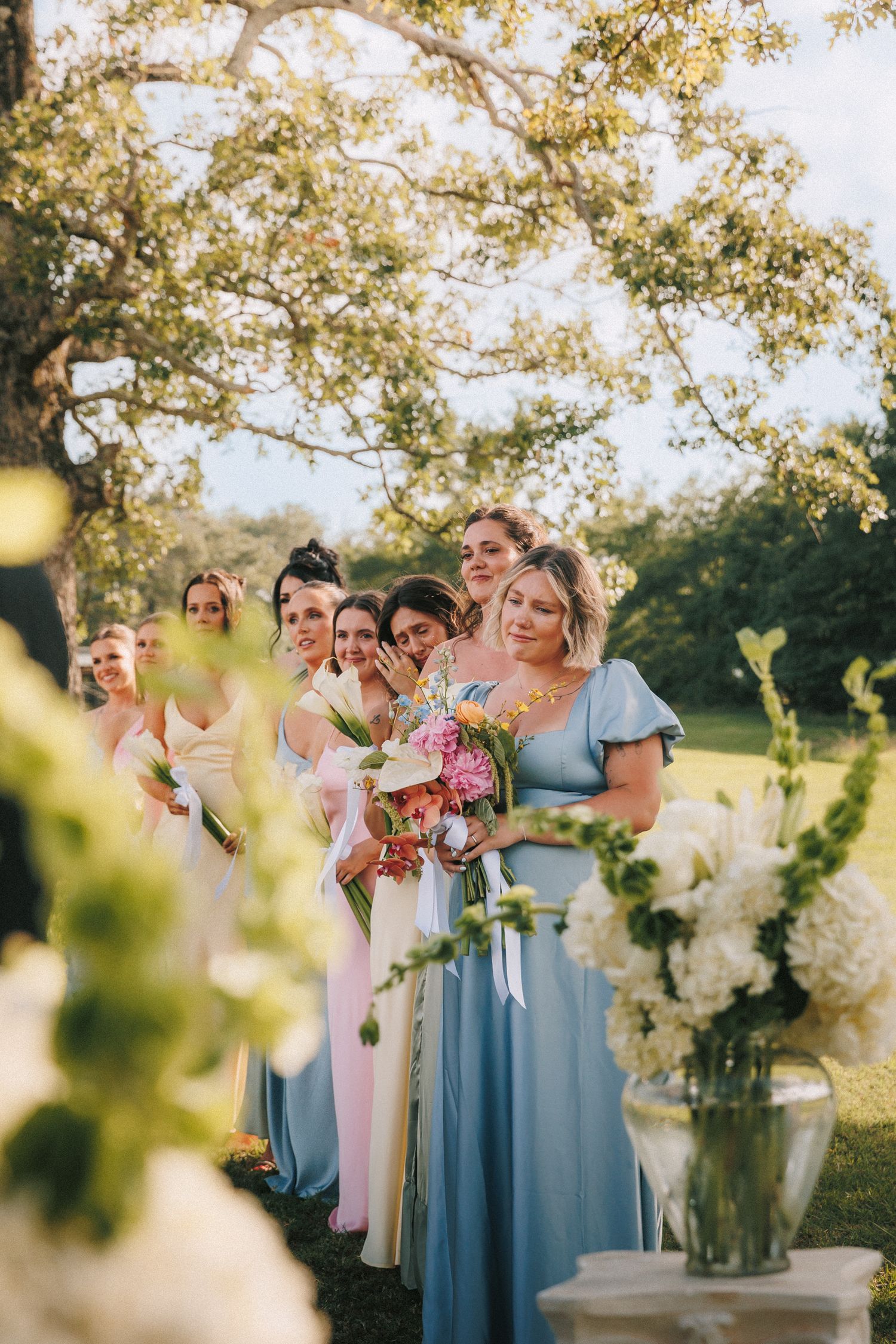Bridesmaids in light blue dresses gather outdoors beside floral arrangements under sunlit trees.