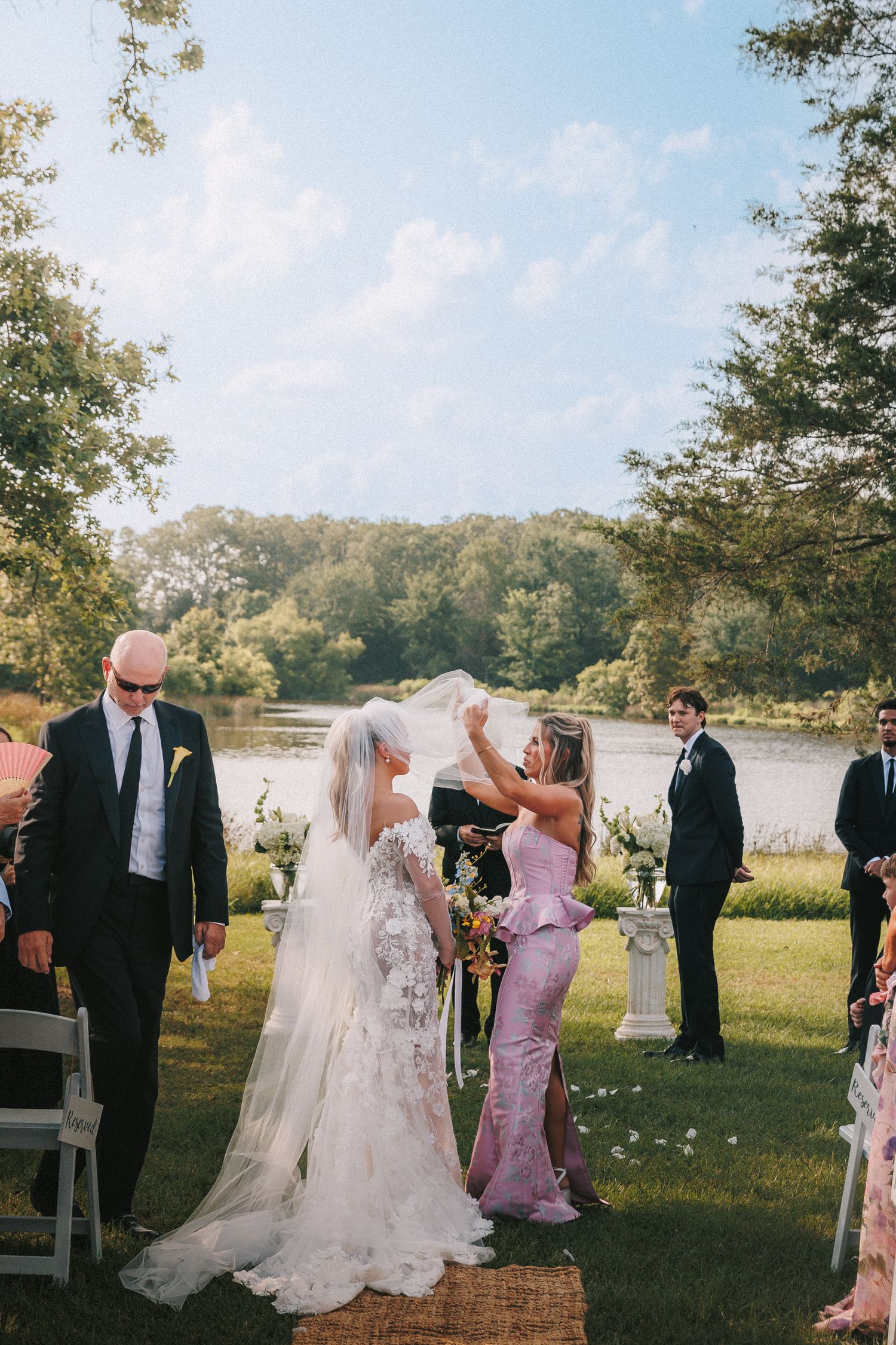 Wedding guests gather in a scenic outdoor setting with trees and a lake during an autumn ceremony.