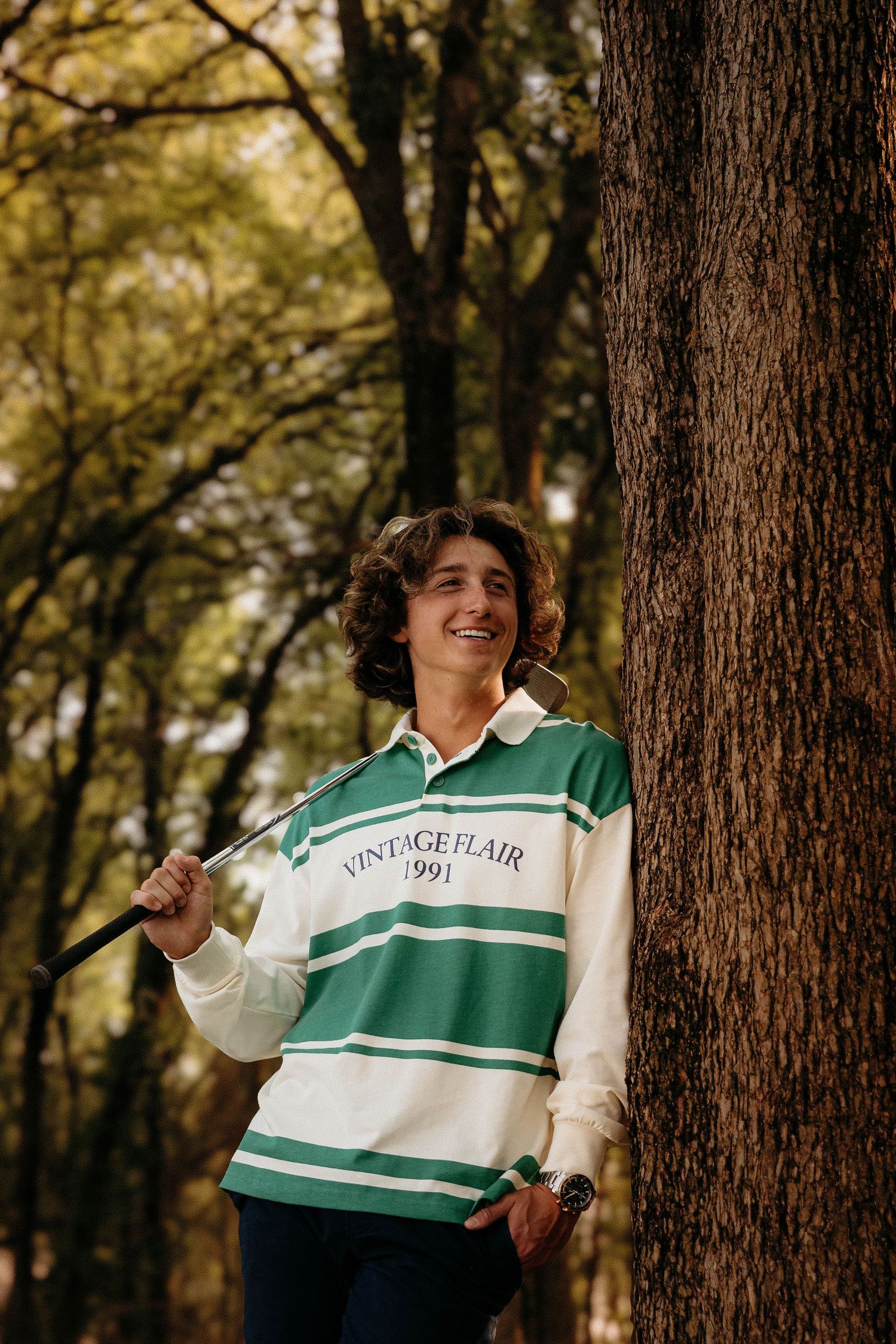 Person posing outdoors in a green and white striped rugby style shirt against a wooded backdrop.