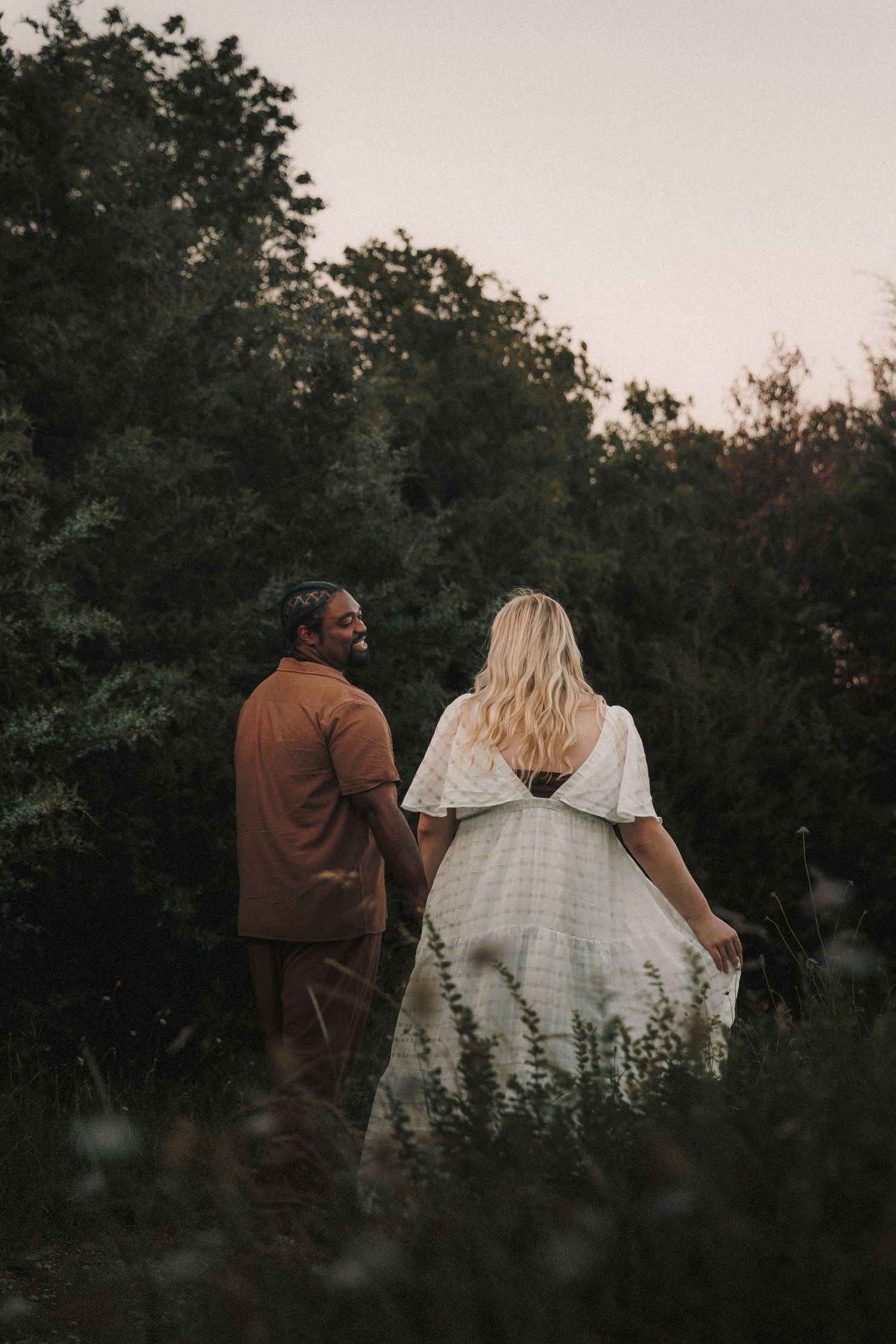 A couple in brown and white attire walking together at dusk with trees silhouetted in the background.