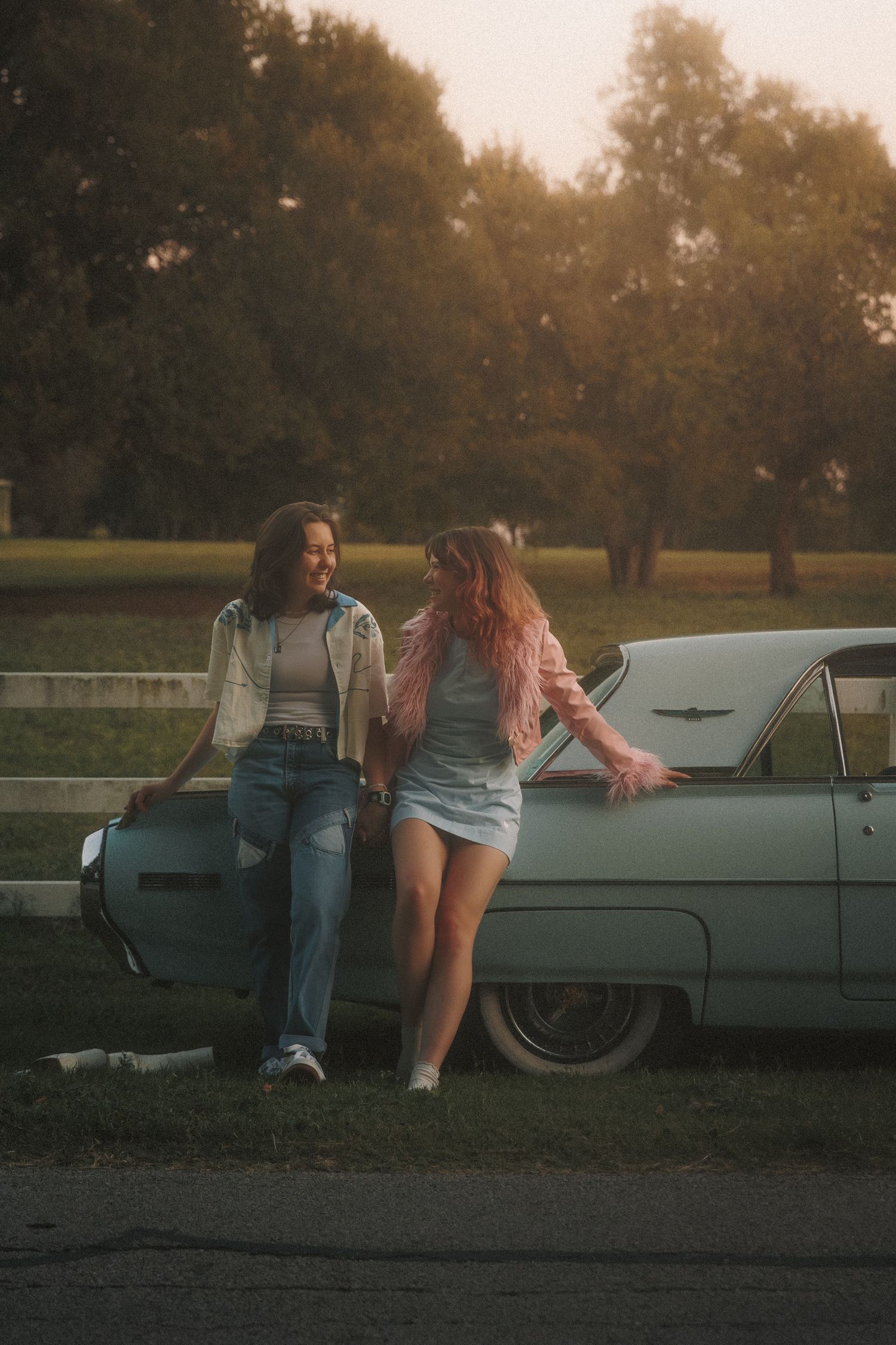 Two friends leaning against vintage car at sunset in park with golden hour lighting.