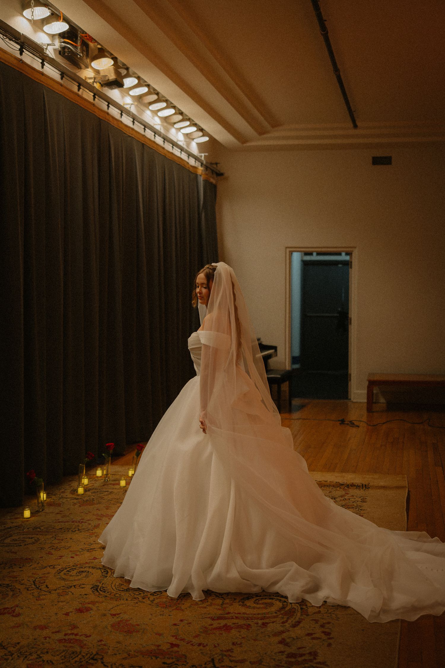 A bride in a flowing white gown and veil stands elegantly in a dimly lit room with candles.