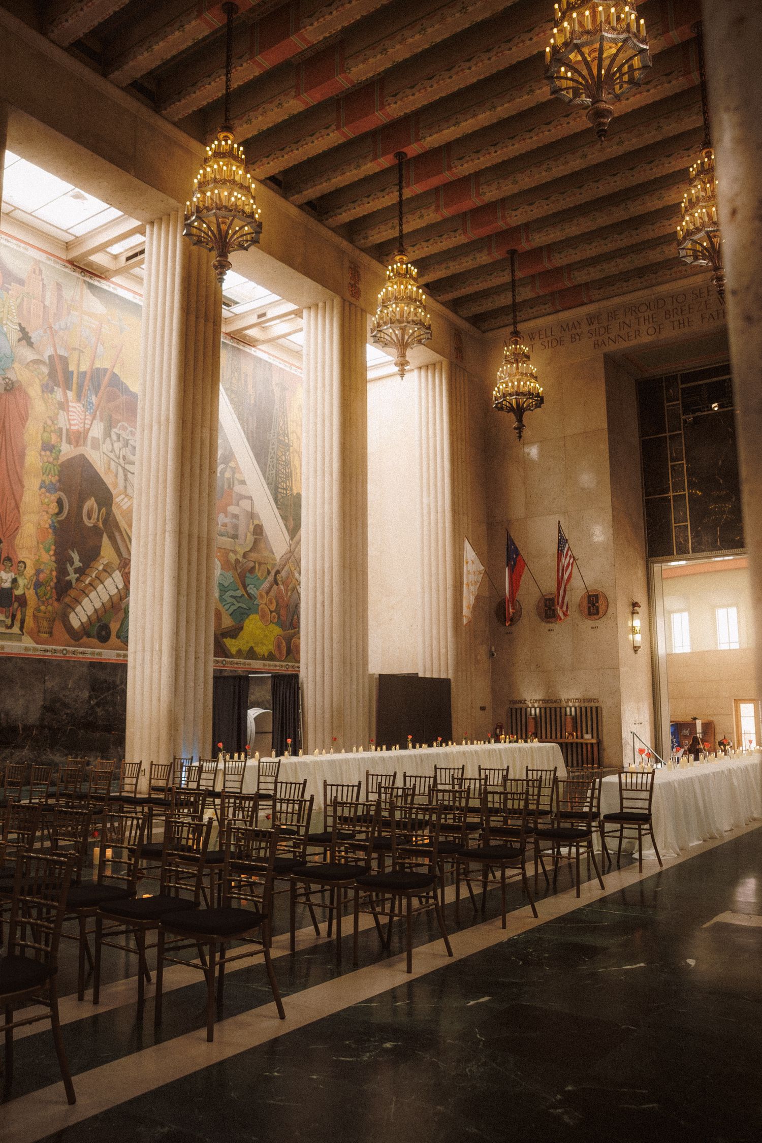 Grand hall with tall columns, chandeliers, rows of chairs, and a long banquet table set for an elegant event.