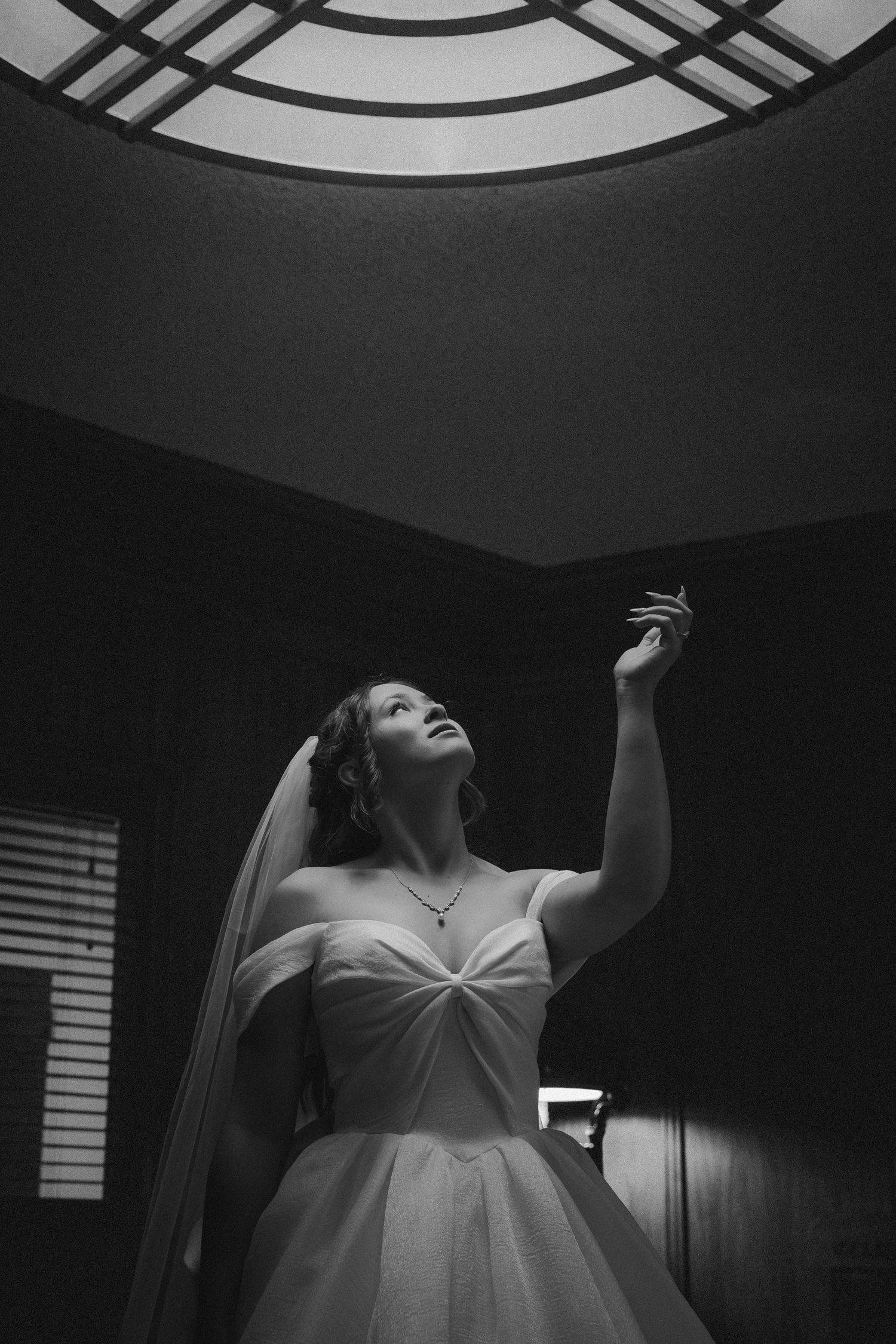 Bride in white gown and veil reaches upward beneath circular skylight in dramatic black and white photo.