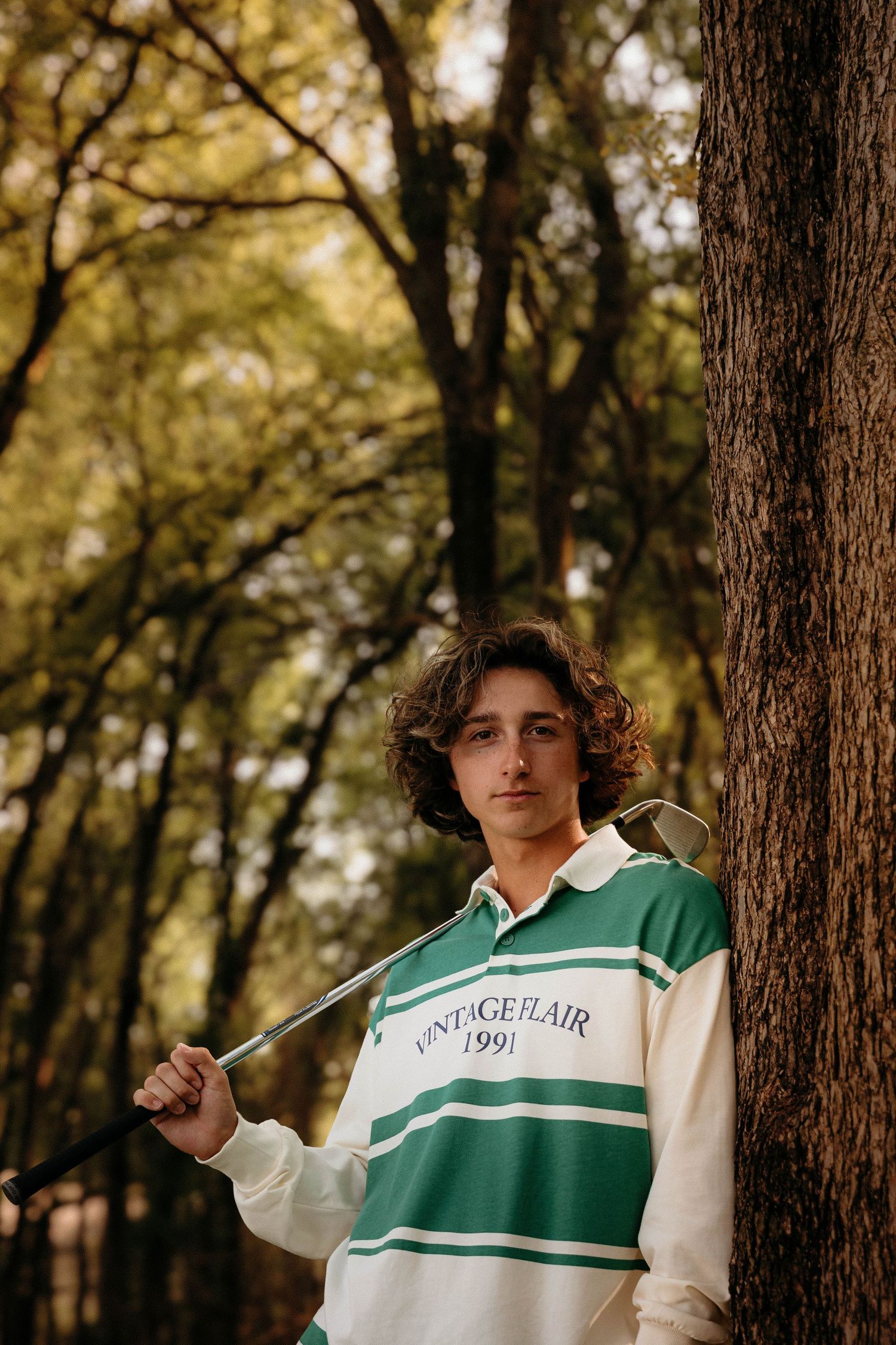 A person wearing a green and white varsity style sweater poses thoughtfully against a wooded forest backdrop.