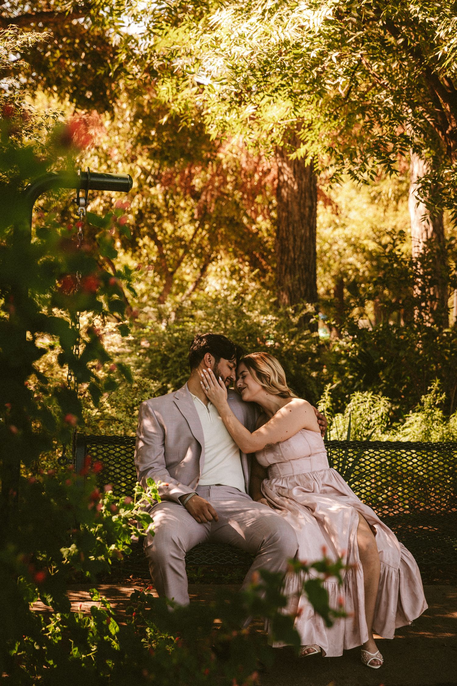 A couple in light-colored attire shares tender moments on a garden bench surrounded by golden sunlight.