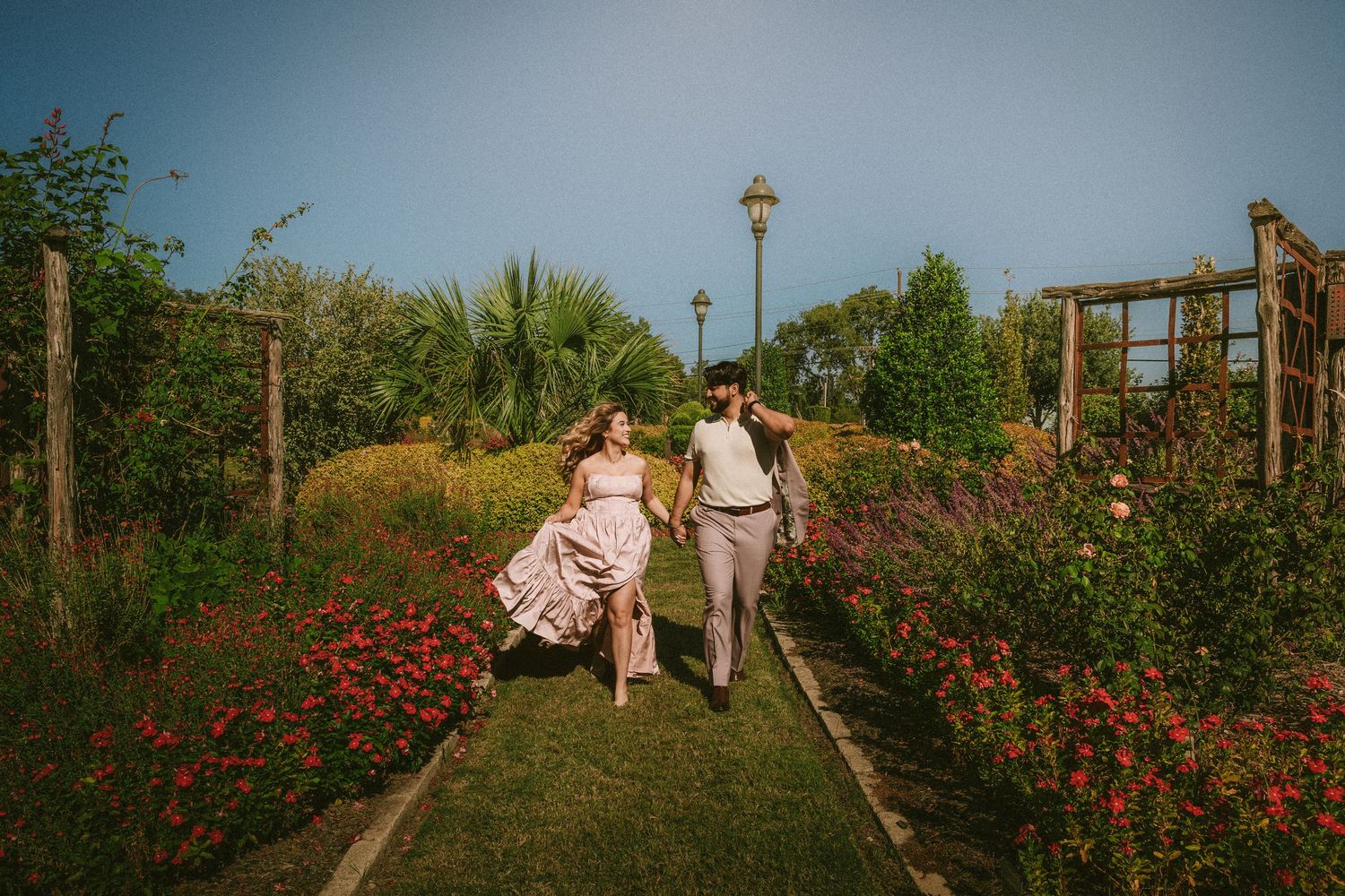 A couple walks through a scenic garden path with blooming red flowers and lamp posts.
