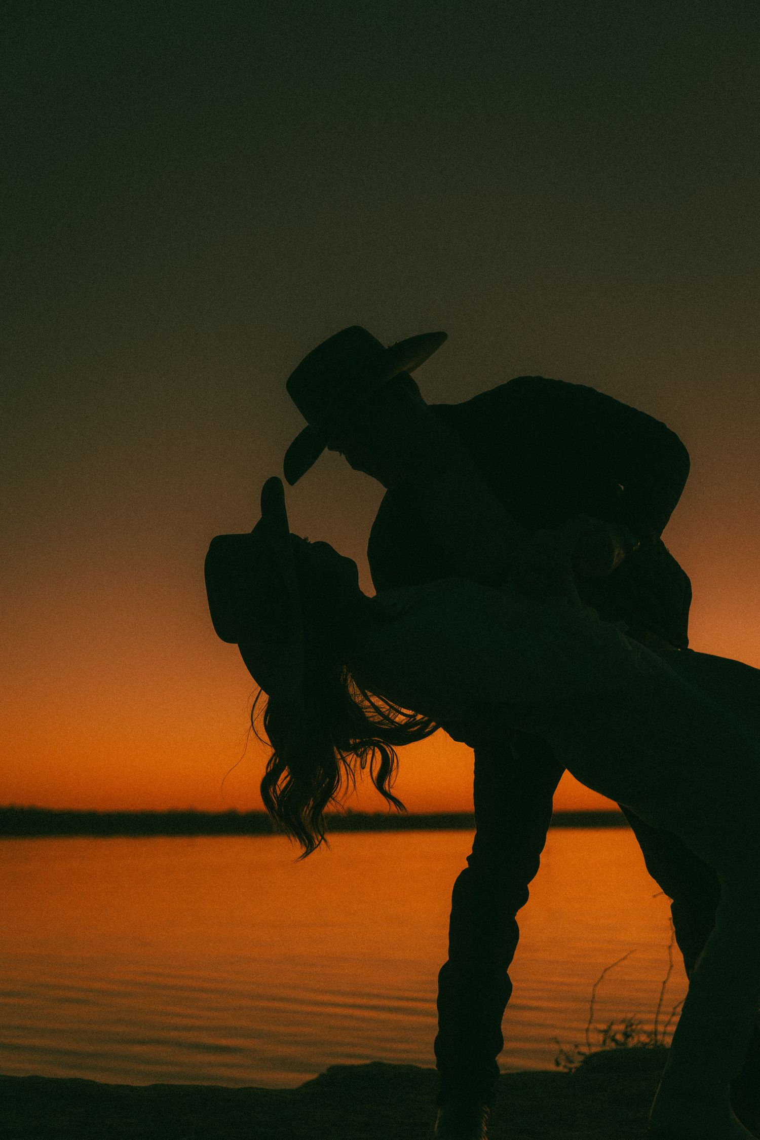 Dramatic silhouette of a couple in a dip pose against an orange sunset sky over water.