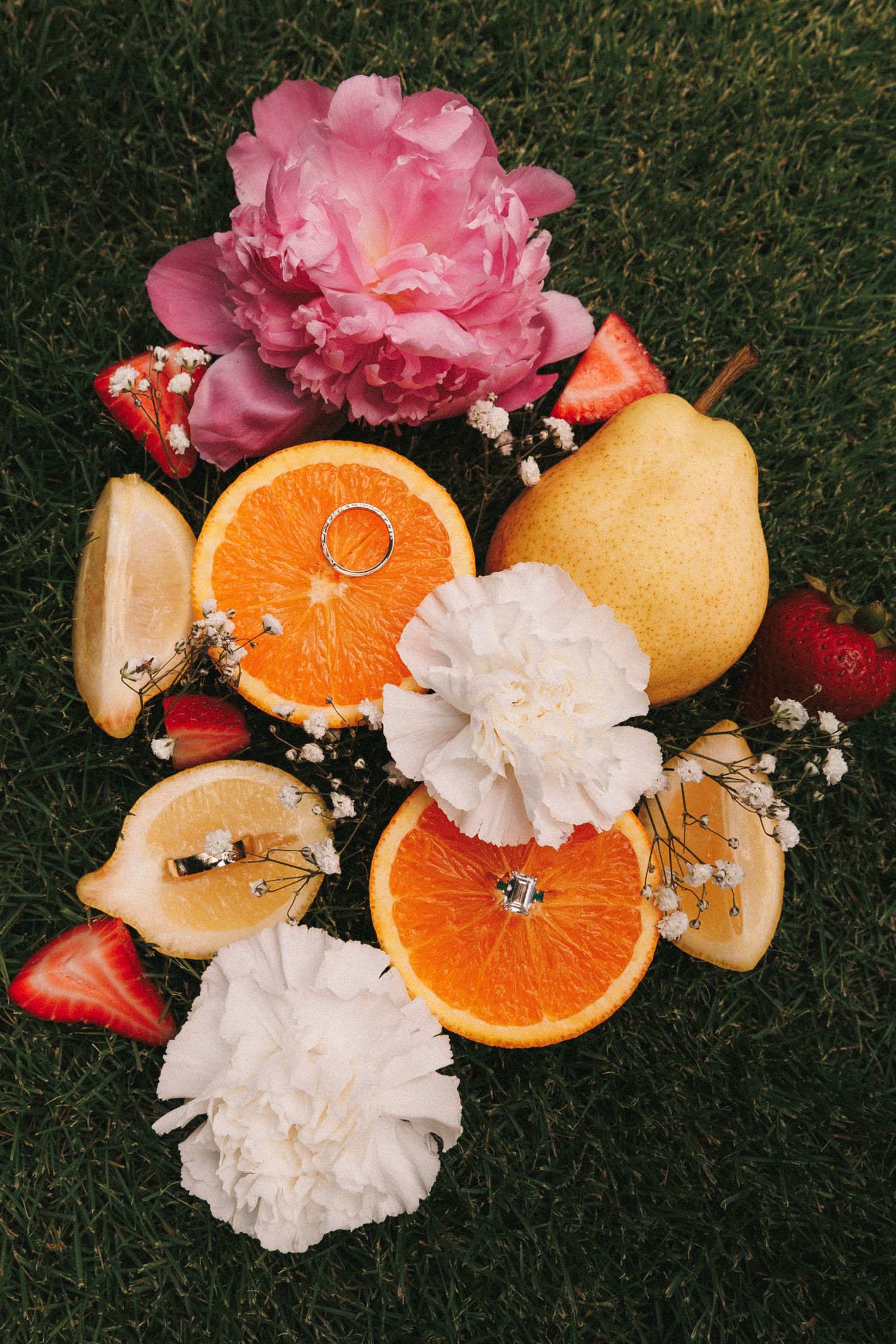 Pink and white carnations arranged with fresh orange slices and fruit on dark grass create a vibrant flat lay composition.