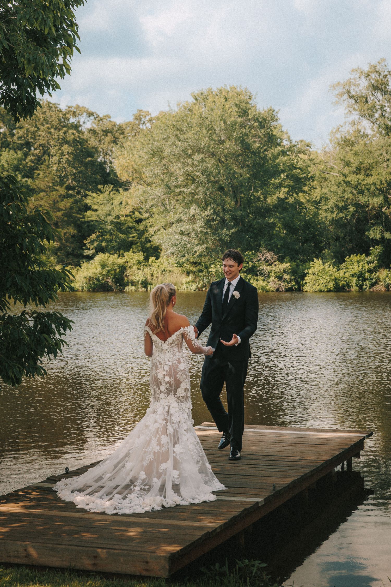 Romantic wedding couple walking together on a wooden dock by the lake with trees in the background.