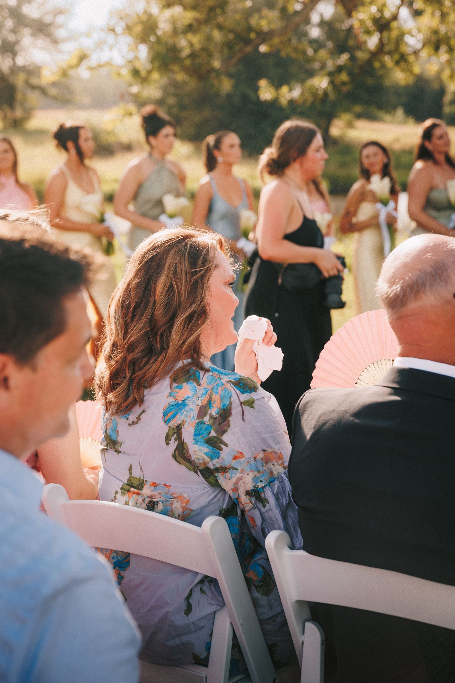 Wedding guests seated in white folding chairs at an outdoor ceremony in warm sunlight.