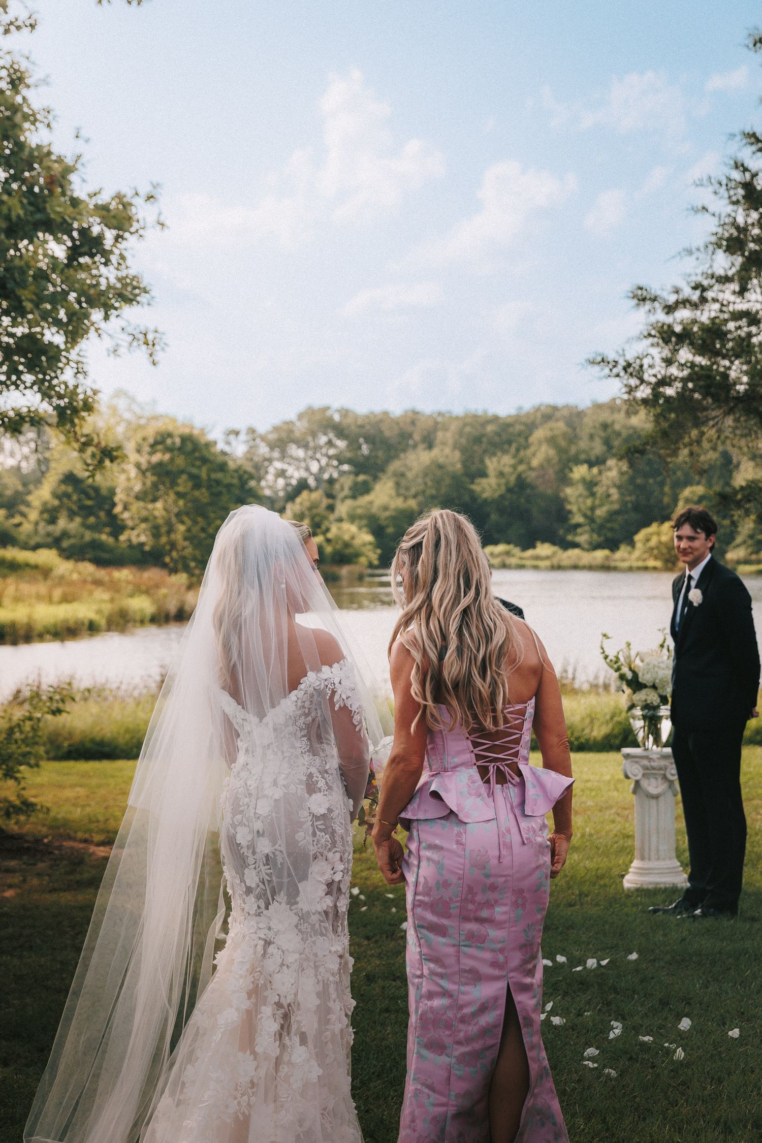 A couple shares an intimate moment on their wedding day by a lake while others look on in the background.
