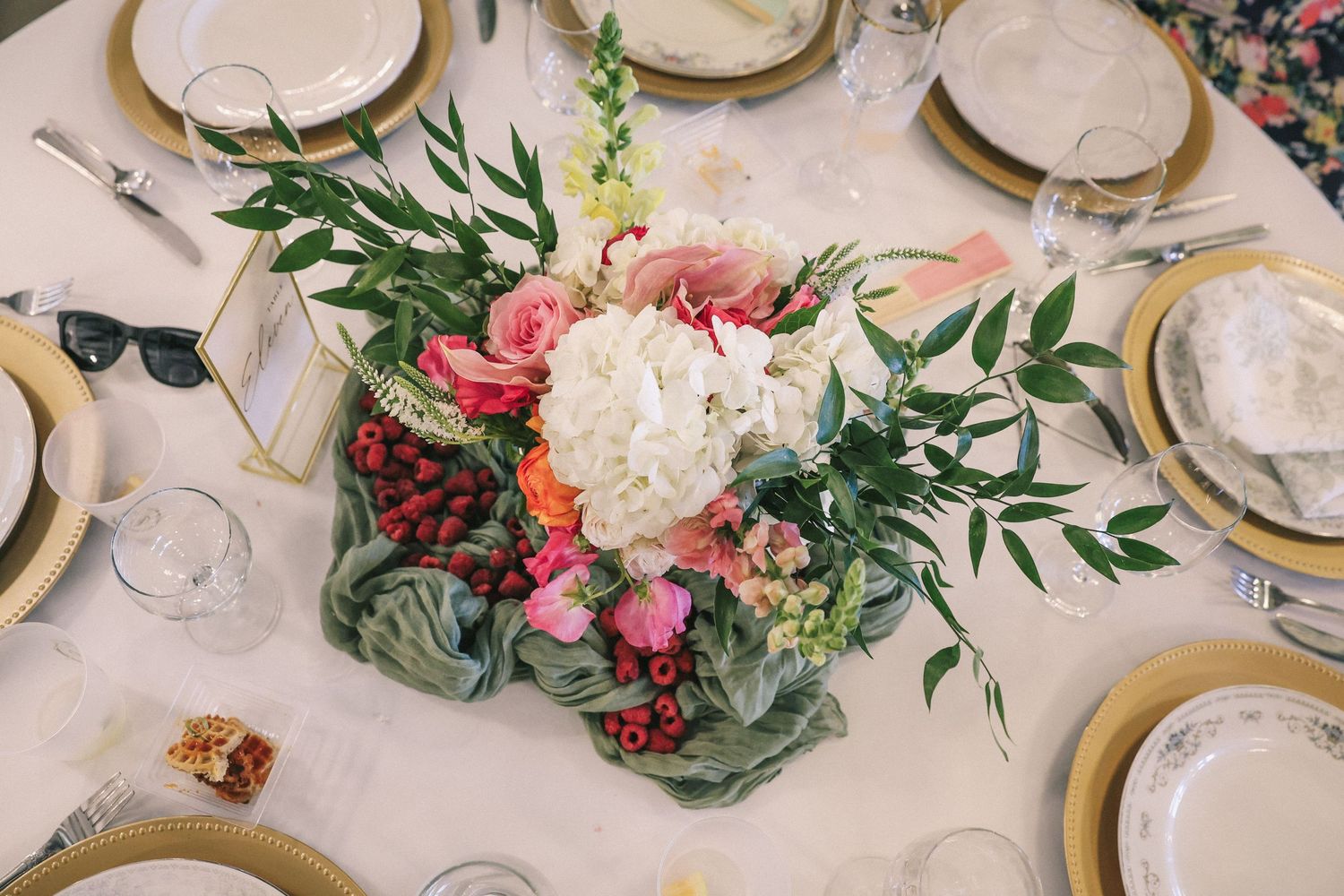 Elegant wedding reception table settings with gold accents, white flowers and greenery on a light tablecloth.