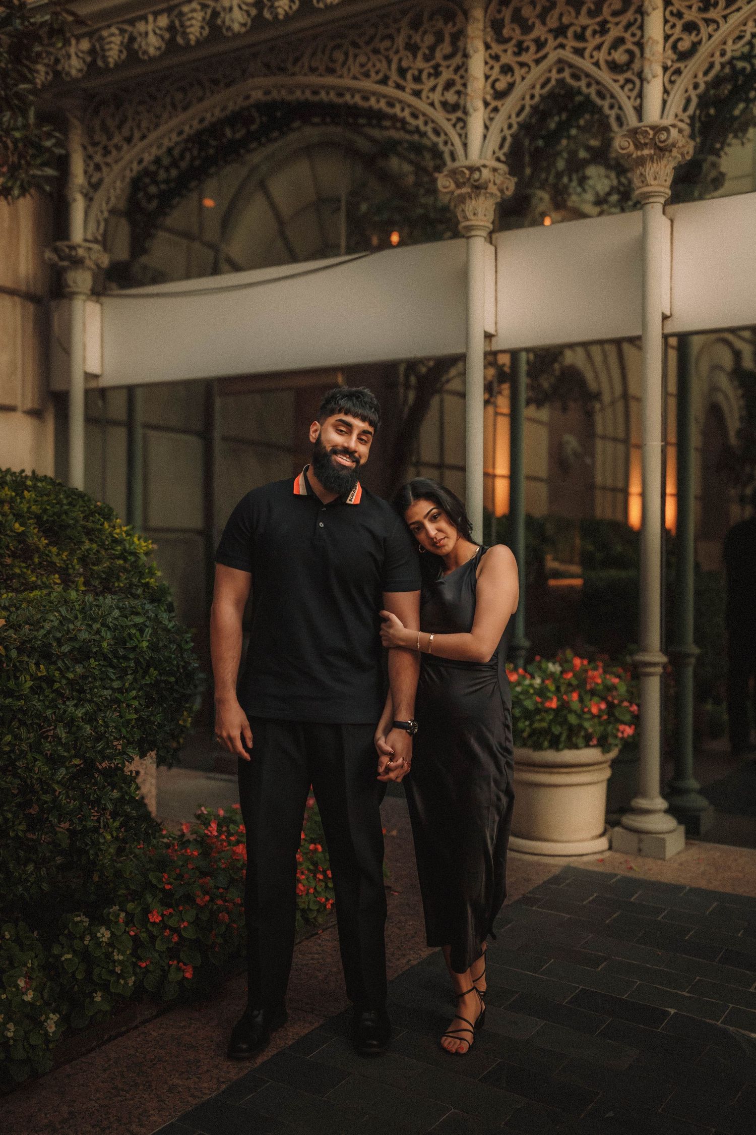 Two people share a romantic moment in an ornate courtyard with decorative columns and floral gardens at dusk.