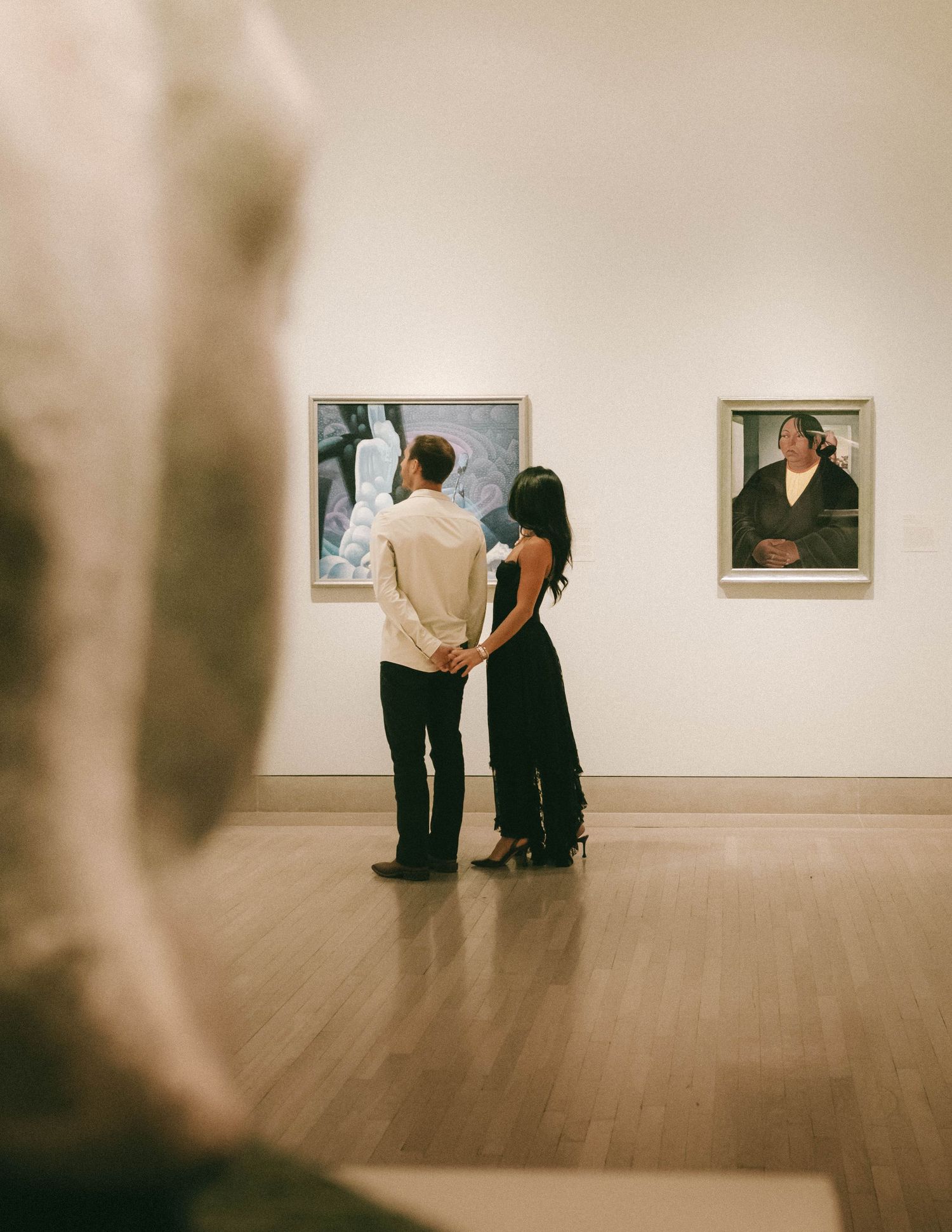 Gallery visitors study artwork displayed on white walls in a minimalist museum space with polished wooden floors.