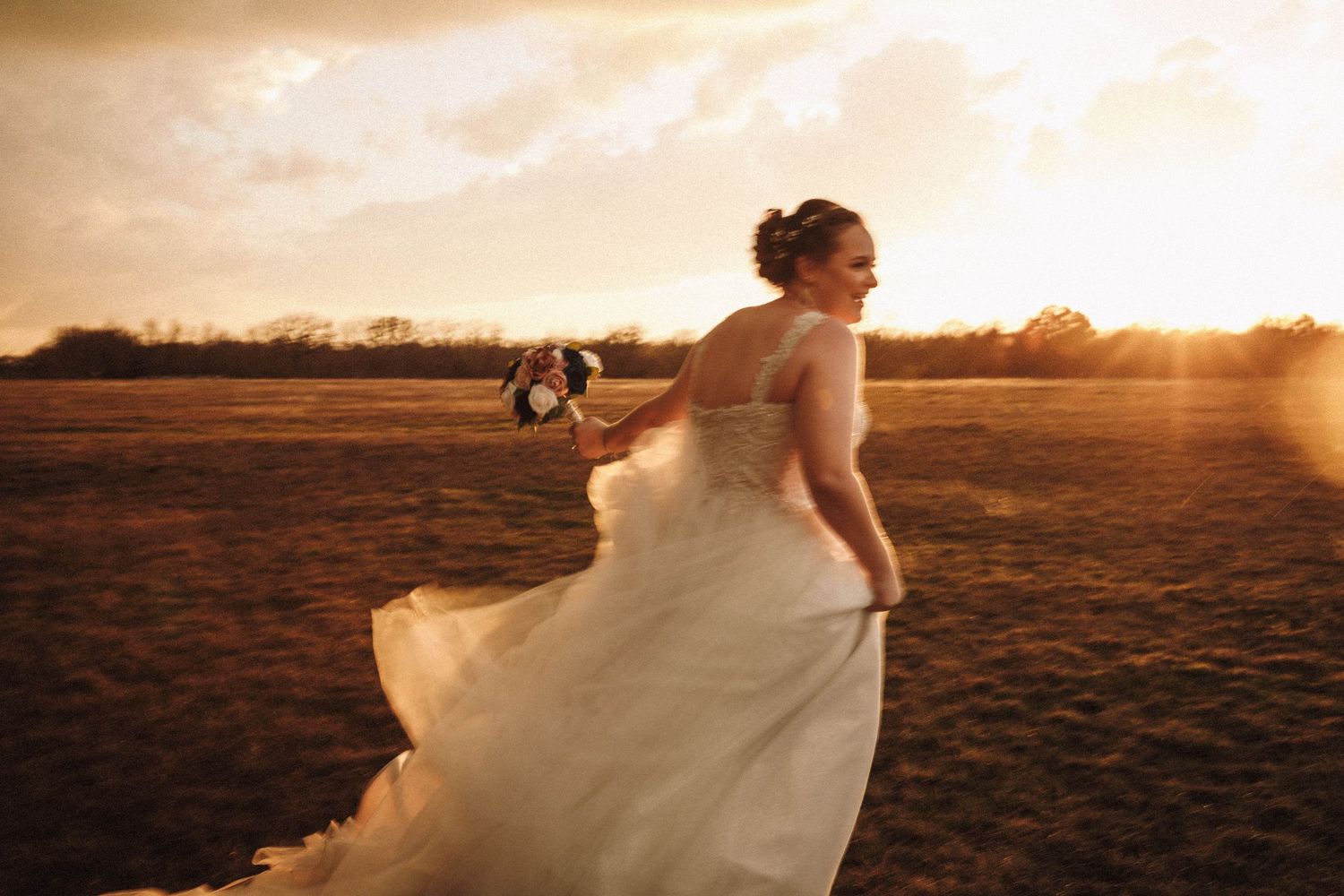 Bride in flowing white gown twirls gracefully in a field at sunset creating a romantic silhouette.