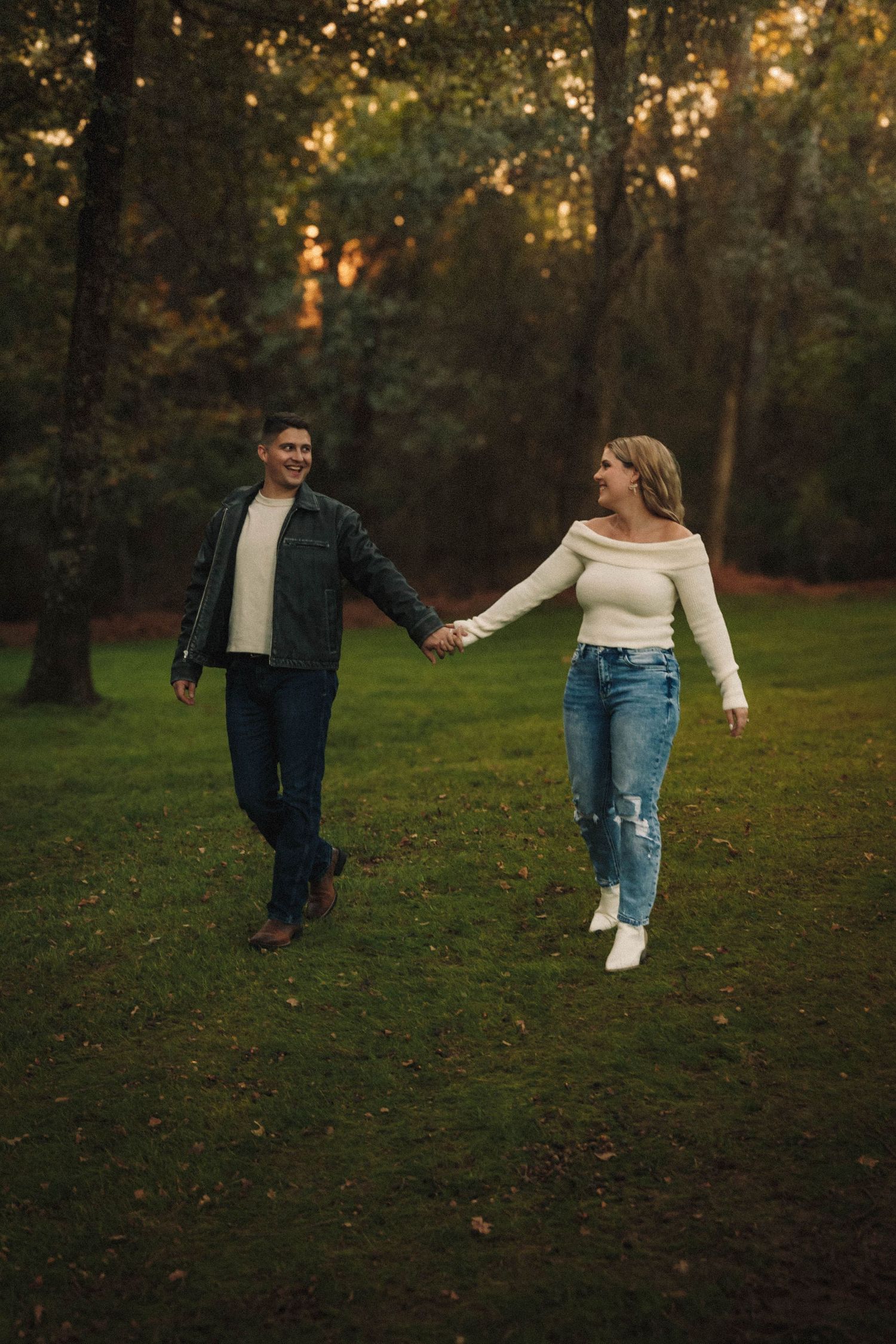 Couple holding hands while walking through a park at golden hour with trees in the background.