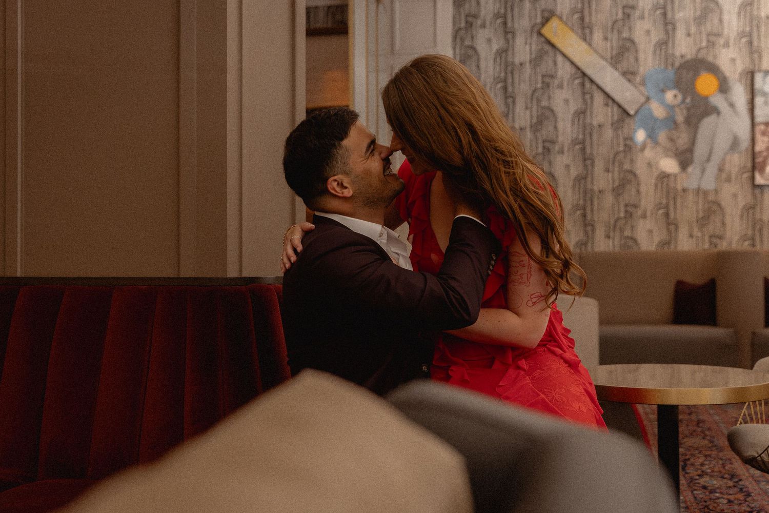 Couple sharing a romantic kiss on a red sofa in an elegant hotel lounge, woman in red dress.