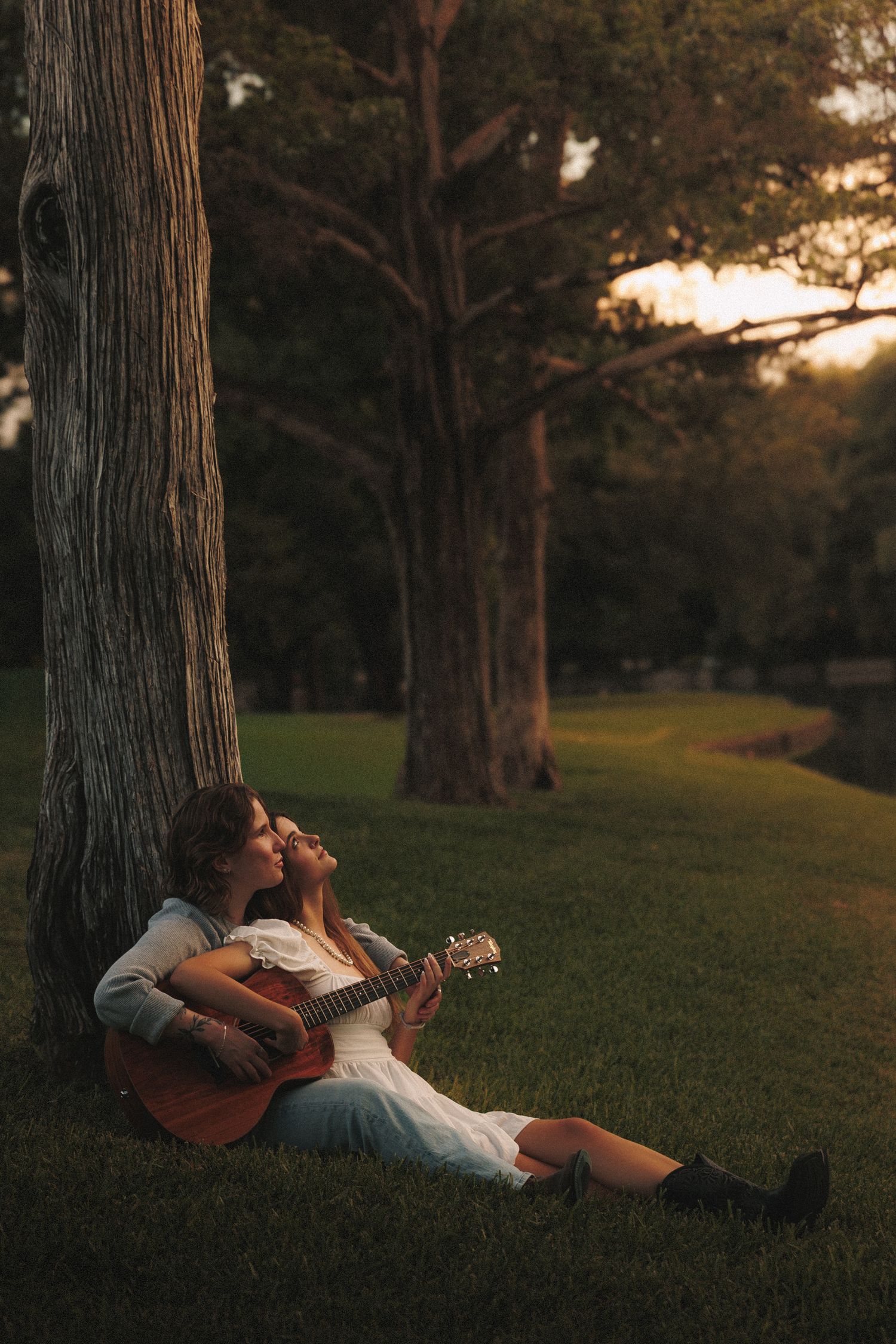 A couple relaxes together on a grassy hillside at sunset while playing acoustic guitar in a peaceful outdoor setting.