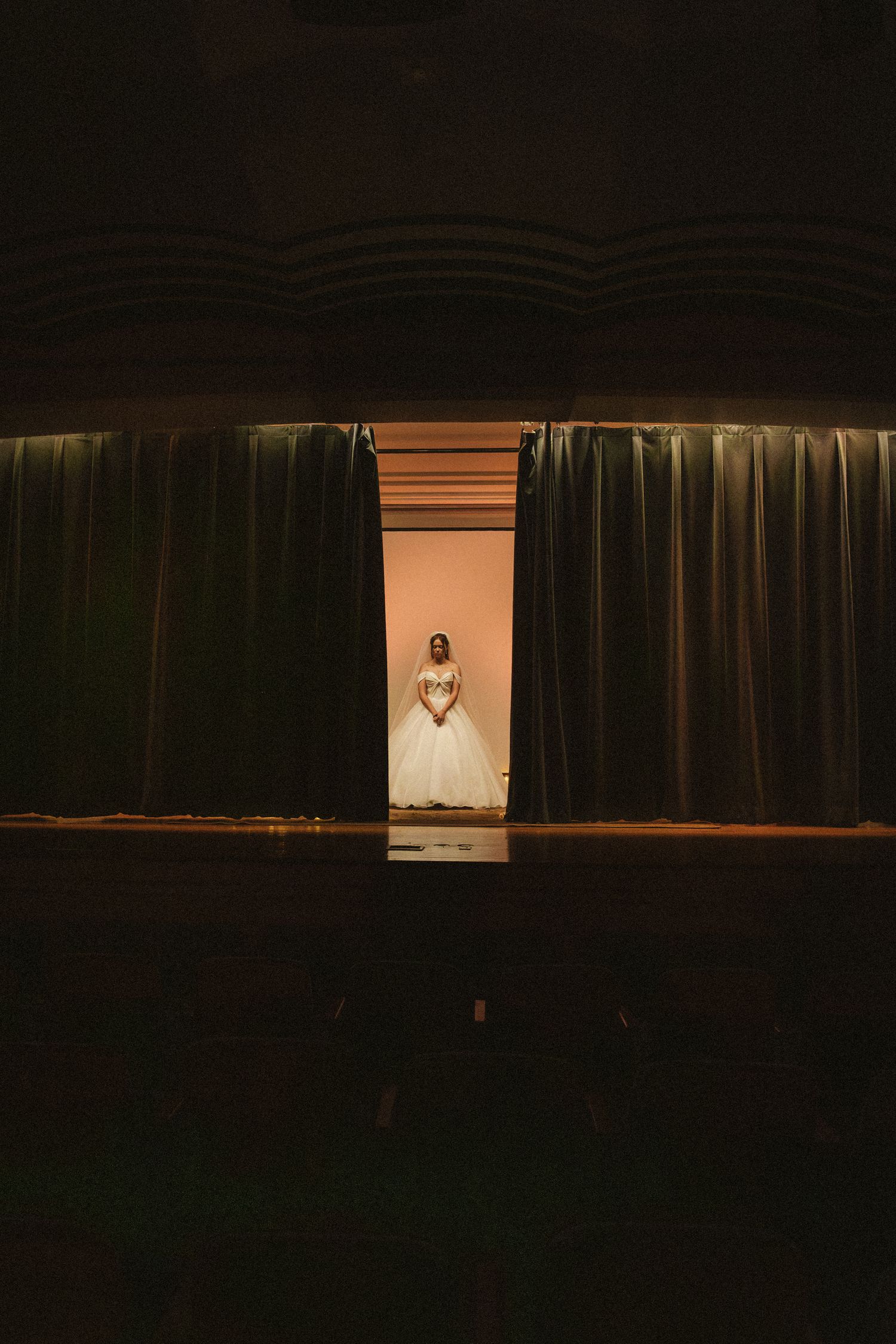 Bride in white ball gown standing in lit doorway framed by dark theater curtains.