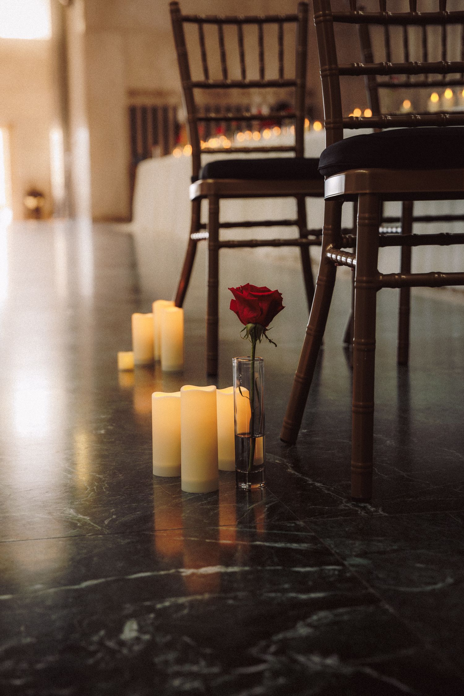 Elegant wedding aisle with white pillar candles and a single red rose in a glass vase on dark marble floor.