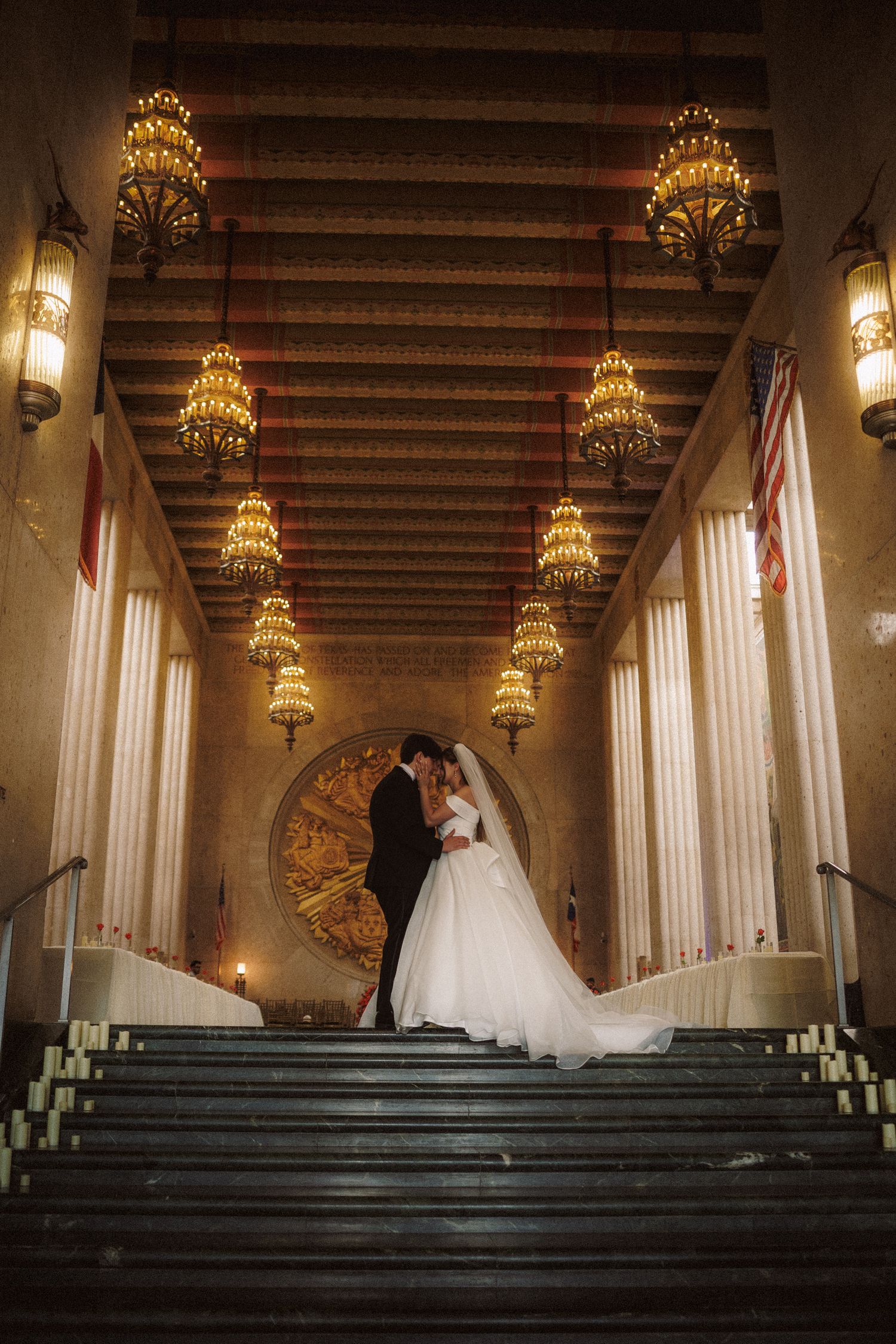 Bride and groom embracing on grand staircase under golden chandeliers in stunning wedding venue.