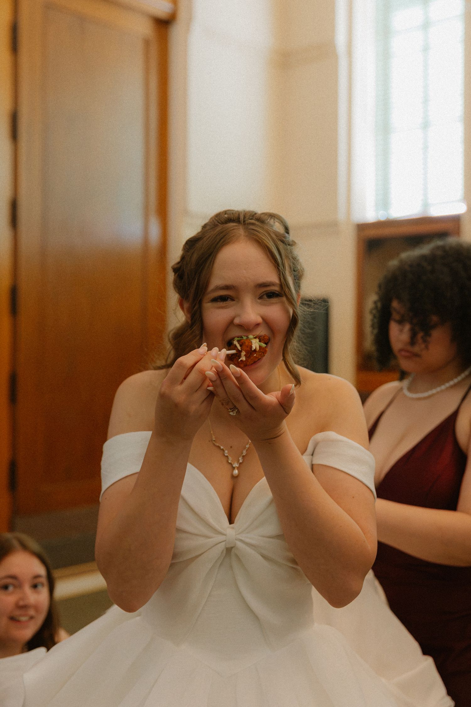 Smiling bride in off-shoulder white gown enjoys wedding cake bite with bridesmaids nearby.